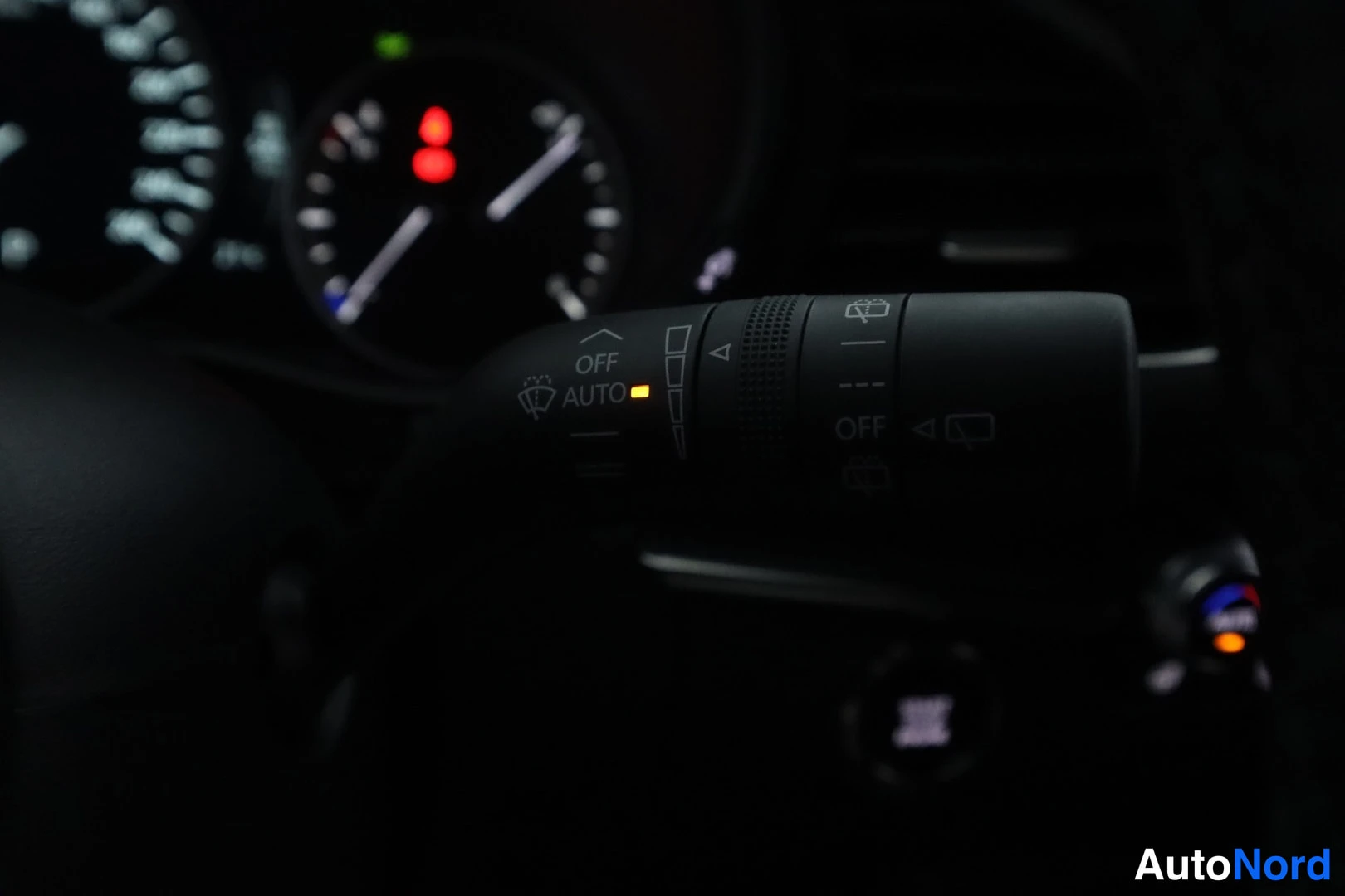 Yksityiskohta: Dark interior close-up of the right-side stalk behind the steering wheel, featuring markings for wiper intensity, auto function, and an illuminated yellow indic