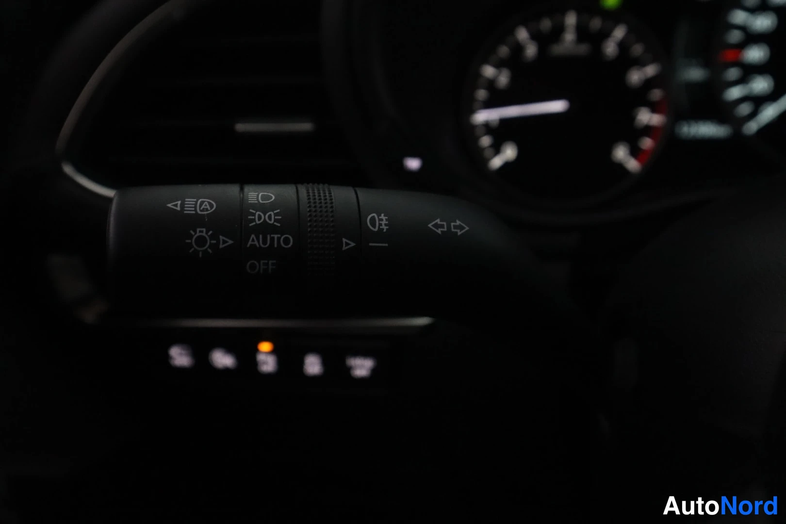 Yksityiskohta: Left-side stalk behind the steering wheel displaying headlight controls, including settings for AUTO operation, low beams, and high beams, set against a dimly l