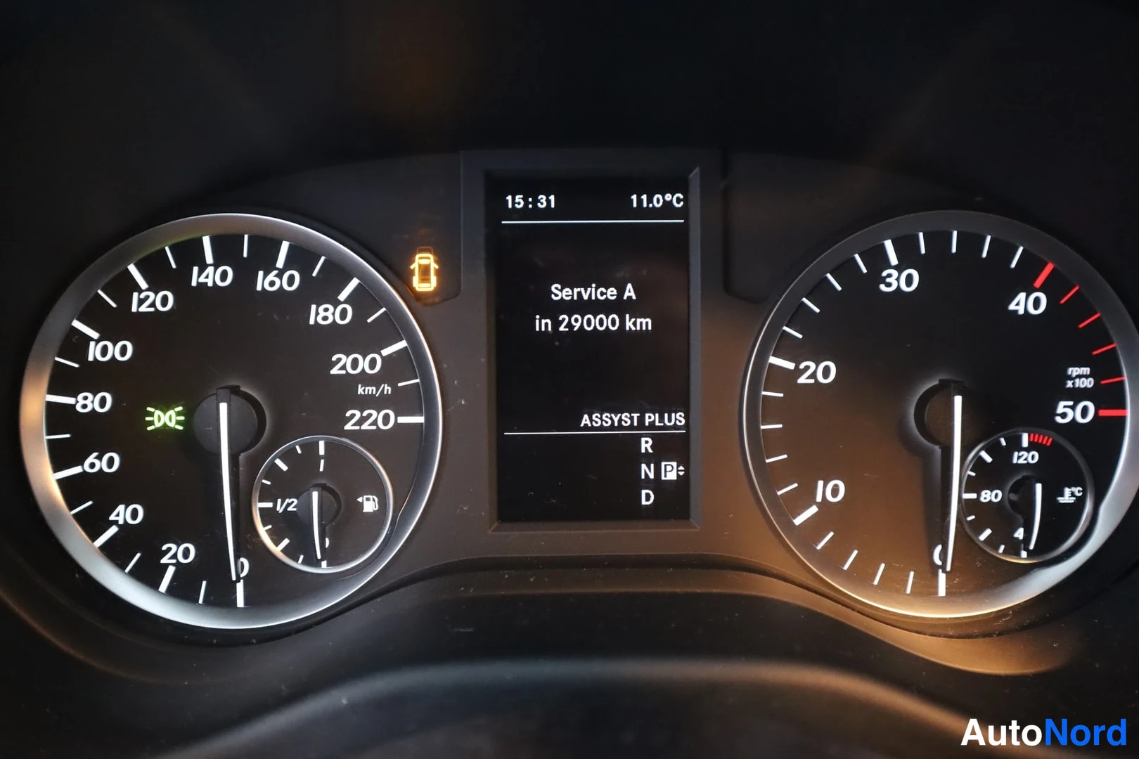 Yksityiskohta: Mercedes-Benzin mittaristo: nopeusmittari asteikolla 0–220 km/h, kierrosmittari asteikolla 0–50 (x100), keskellä näyttö jossa teksti 'Service A in 29000 km' ja