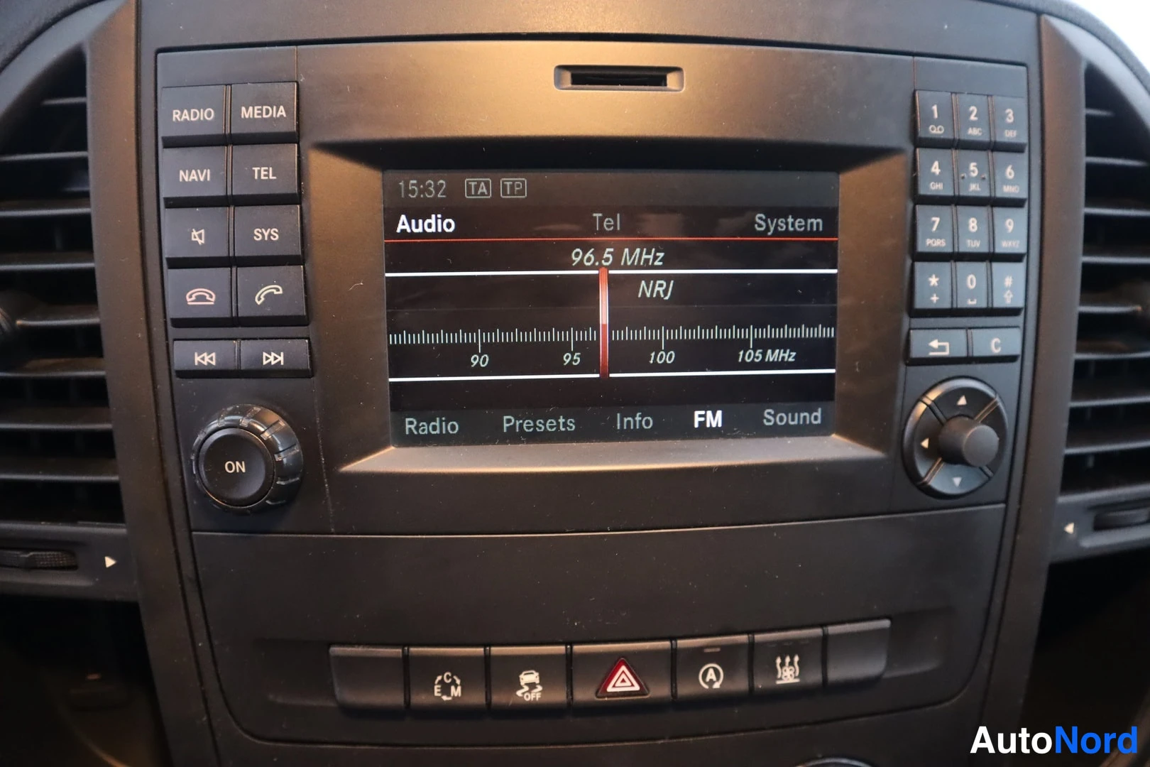 Yksityiskohta: Mercedes-Benzin kojelaudan radionyksikkö, näytössä Audio/Tel/System-valikot ja FM-taajuus 96,5 MHz 'NRJ', ympärillä RADIO/MEDIA/NAVI/TEL-painikkeet ja numeronäp