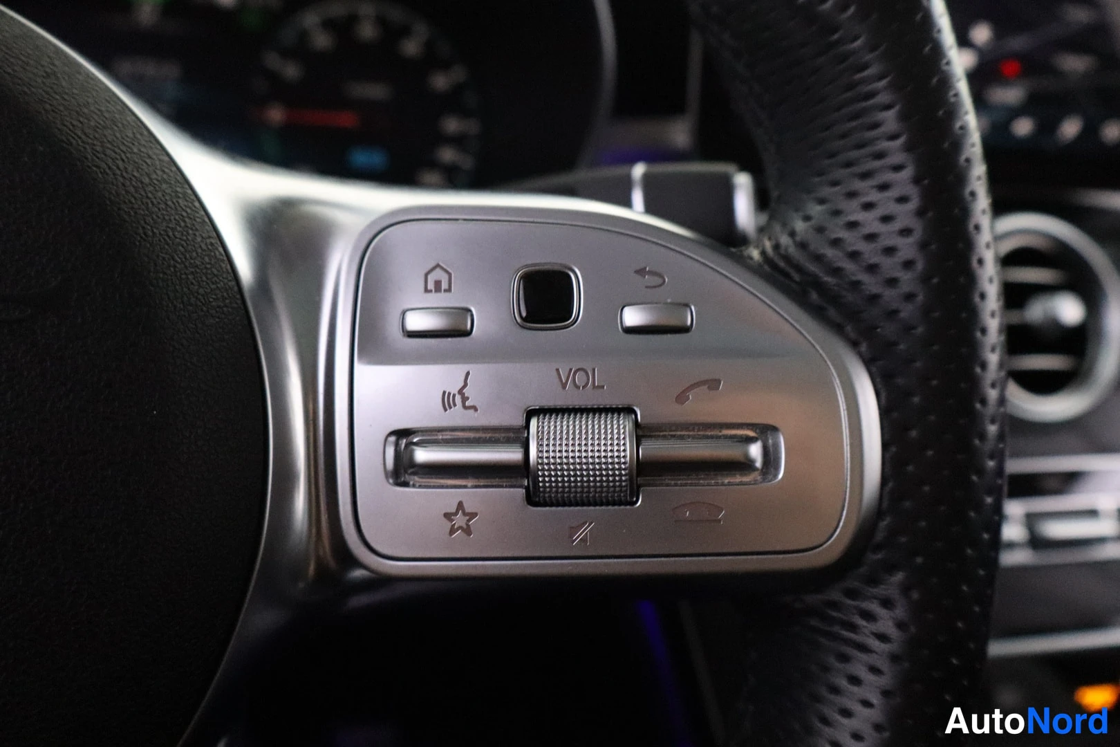 Yksityiskohta: Multifunction steering wheel control on the right spoke with silver trim and black perforated leather surround. Buttons include home, back/return, volume knob m