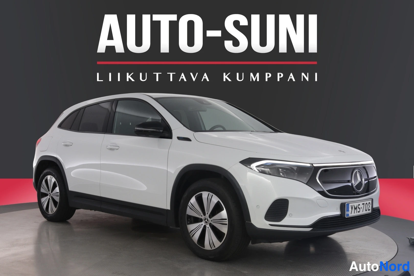 Etuviisto, nokka oikealle: Valkoinen Mercedes-Benz EQA -maastauto etukulmasta autonäyttelylavalla, rekisterikilpi.