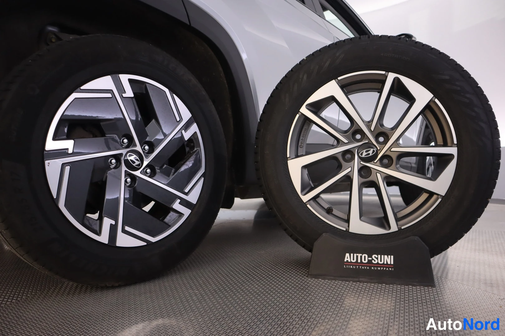 Yksityiskohta: Kaksi Hyundai-kevytmetallivannetta sivittain: autoon asennettu tumma vanne vasemmalla ja irtonainen kaksivärinen vanne oikealla.