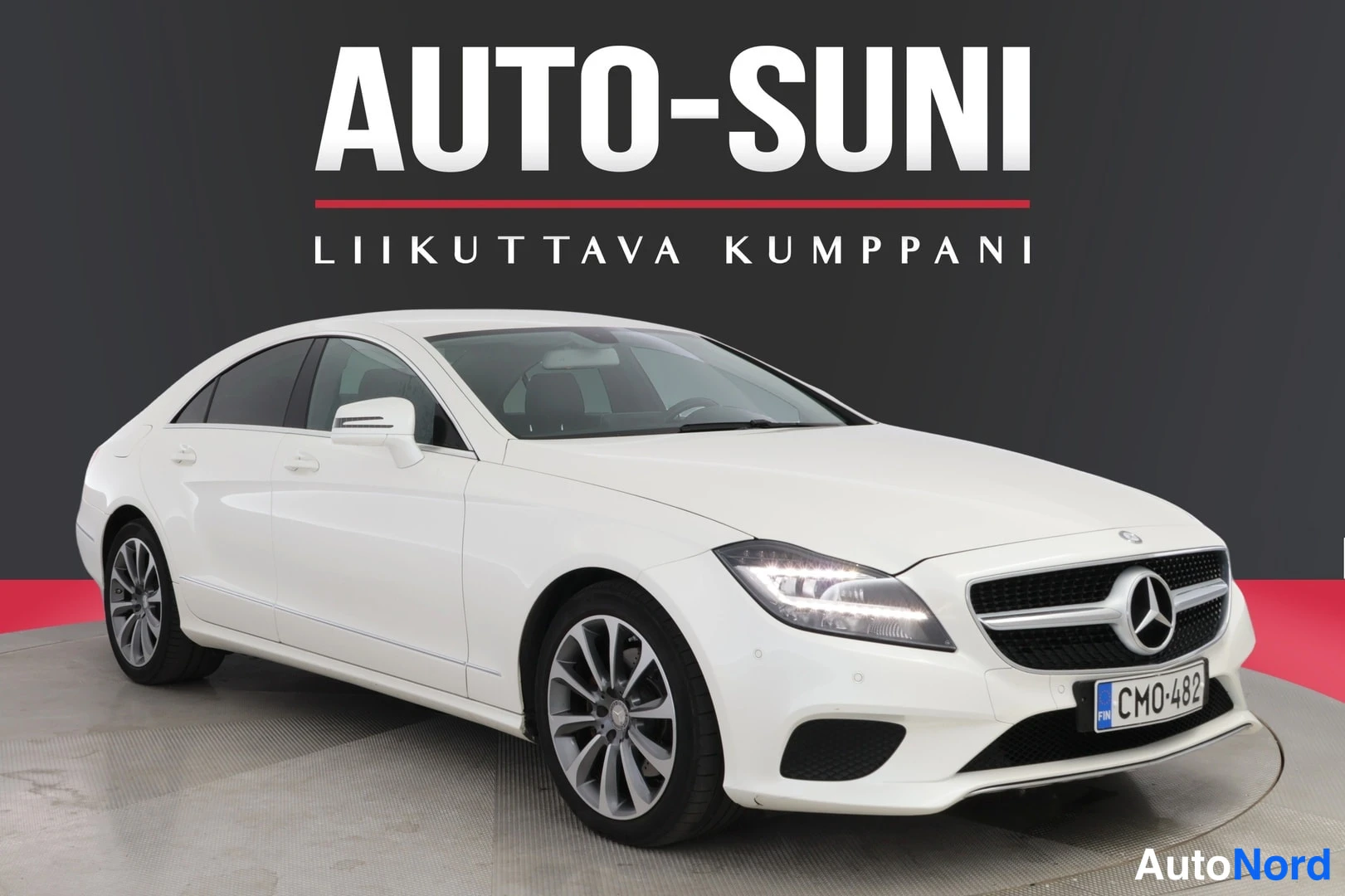 Etuviisto, nokka oikealle: Front three-quarter left view of a white Mercedes-Benz CLS sedan in a showroom. The image displays the front grille with the Mercedes emblem, headlight