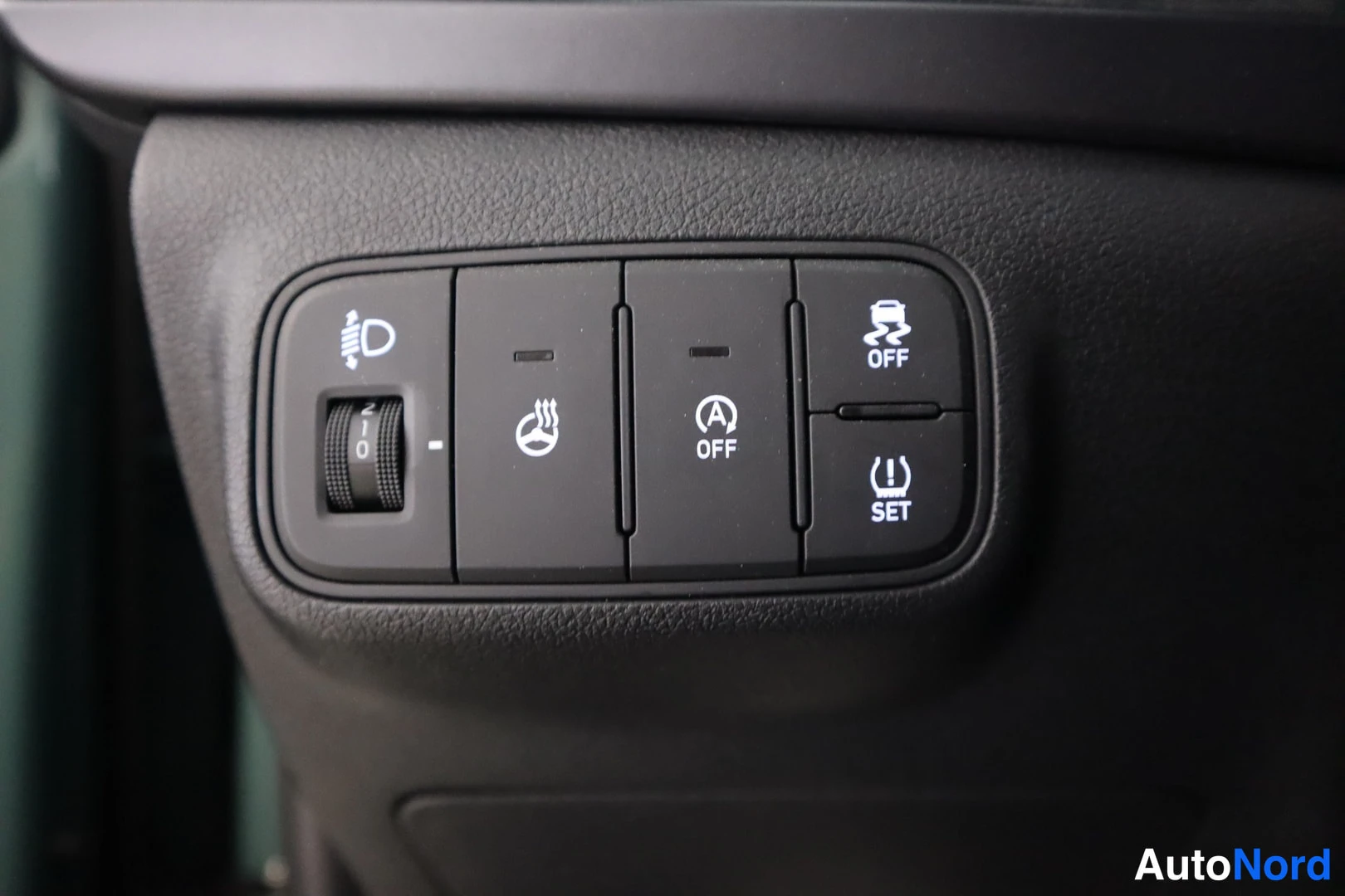 Yksityiskohta: Close-up of dashboard button panel showing headlight rotation dial, heated steering wheel button, auto start-stop off button, and tyre pressure set button.