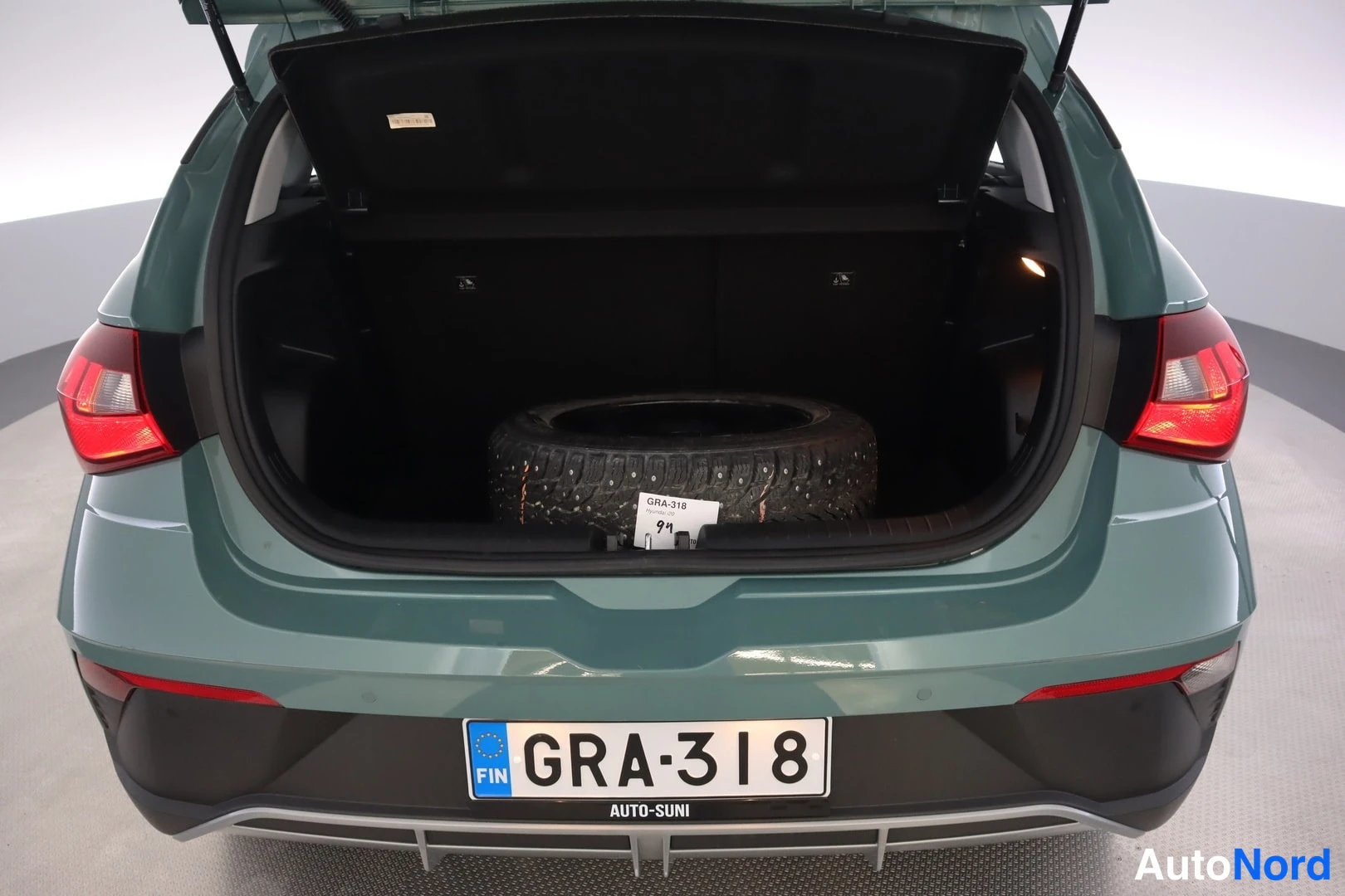 Takaa: Rear view of an open hatchback boot containing a spare winter tyre. The vehicle body is a metallic teal green colour. The rear number plate reads with FIN flag.