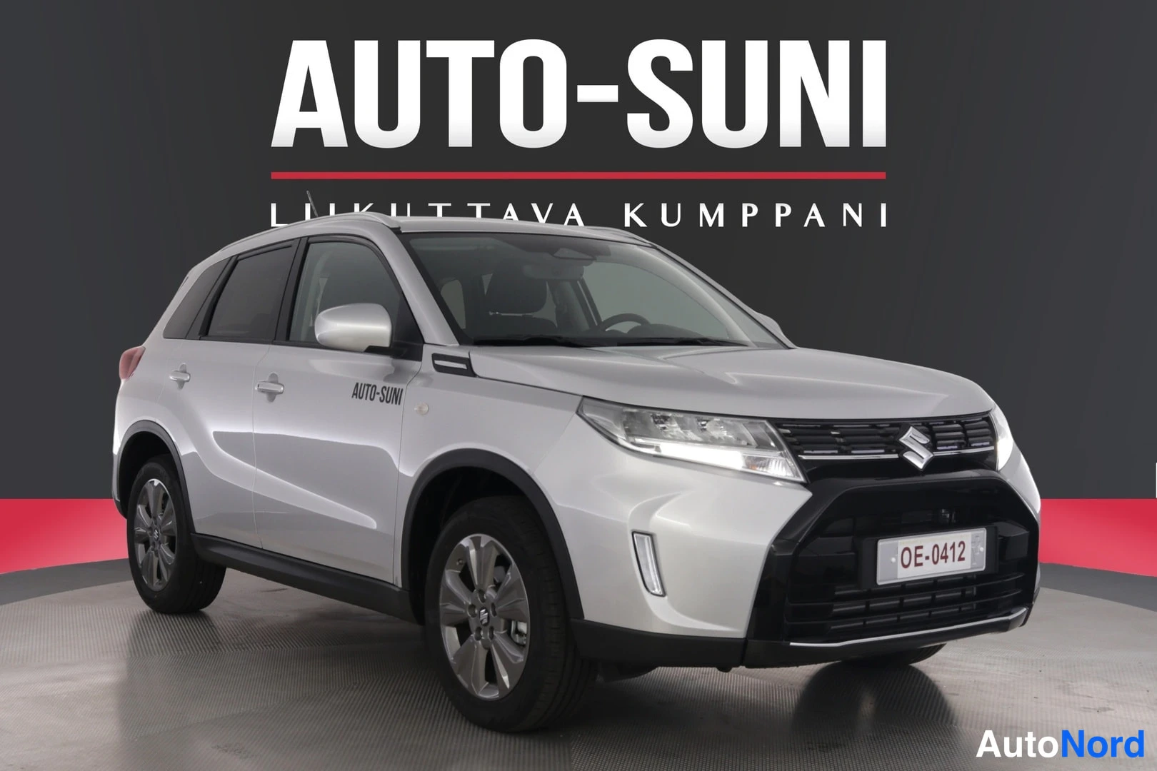 Etuviisto, nokka oikealle: Etusarjakuva valkoisesta Suzuki-kirjaautoista, jossa näkyvät etuosan grilli ja valot sekä vasen sivu. Auto on sisätiloissa, taustalla AUTO-SUNI mainoslog