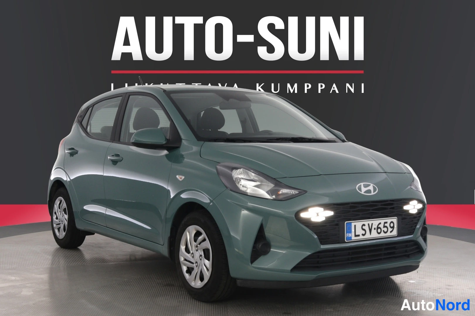 Etuviisto, nokka oikealle: Hyundai i10 -vihreä metallivärinen hatchback, edestä ja vasemmalta kulmauskuvana. Autossa on LED-ajovalot ja takalaudalla rekisterinumero. Auto seisoo gr