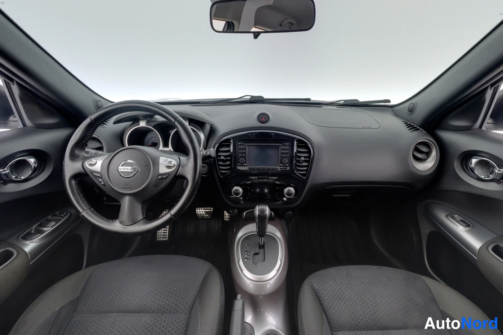 Muu: Sisätila kuvattu kojtaulusta: Nissan-ohjauspyörä, keskikonsolissa näyttö ja automaattivaihteen valitsin, tummat istuimet.