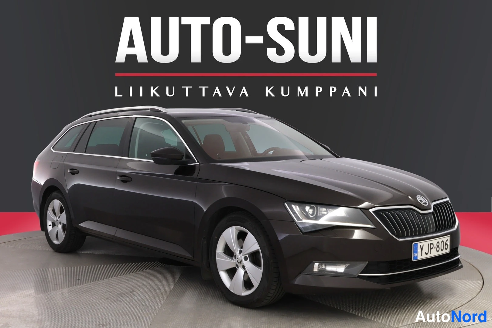 Etuviisto, nokka oikealle: Front three-quarter view of a dark grey Skoda Superb estate car. The grille and headlights are visible along with the front left door and wheel.