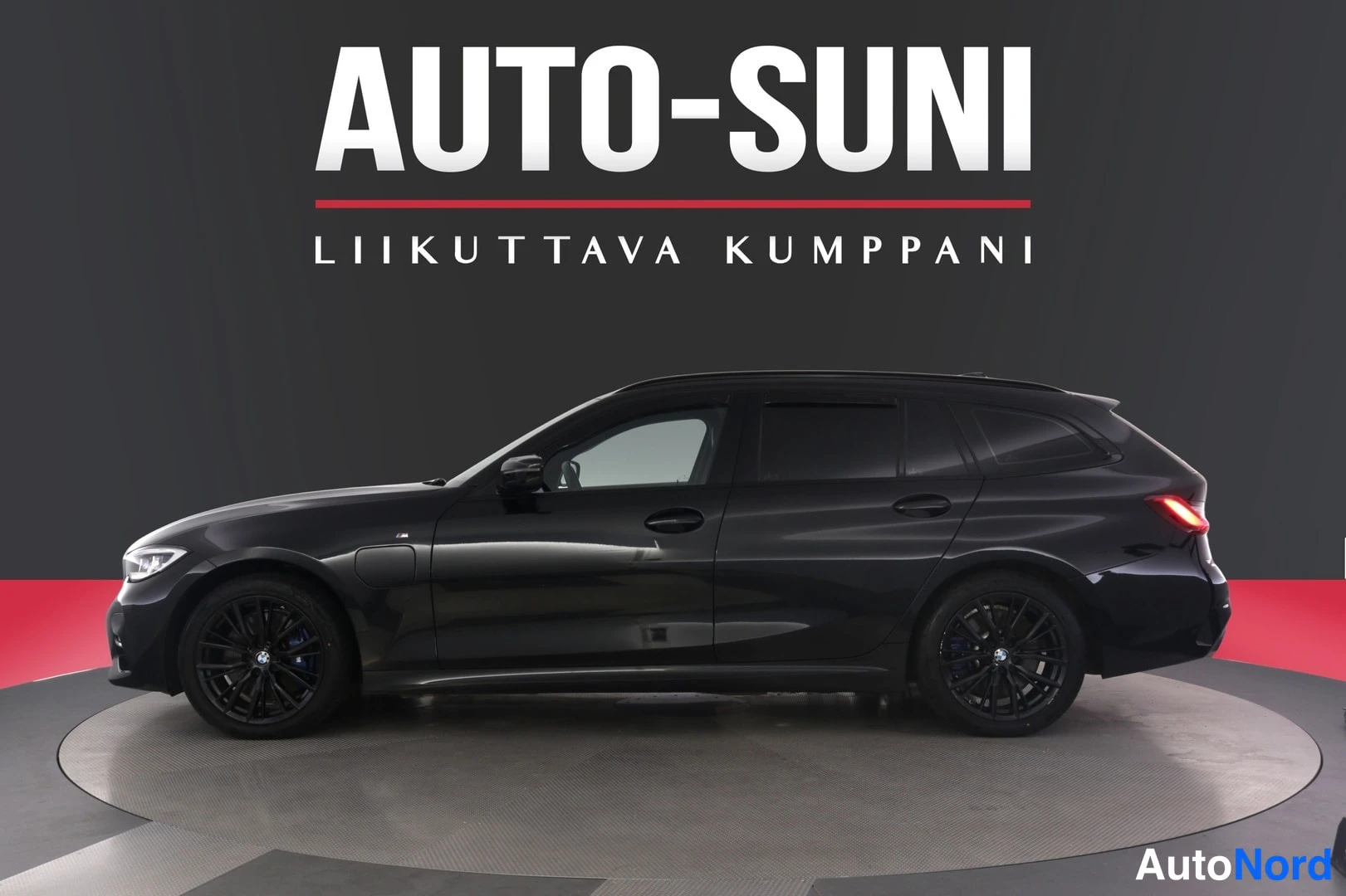 Etuviisto, nokka vasemmalle: Mustan farmariauton sivuprofiili studioalustalla, taustalla AUTO-SUNI-mainoskyltti.