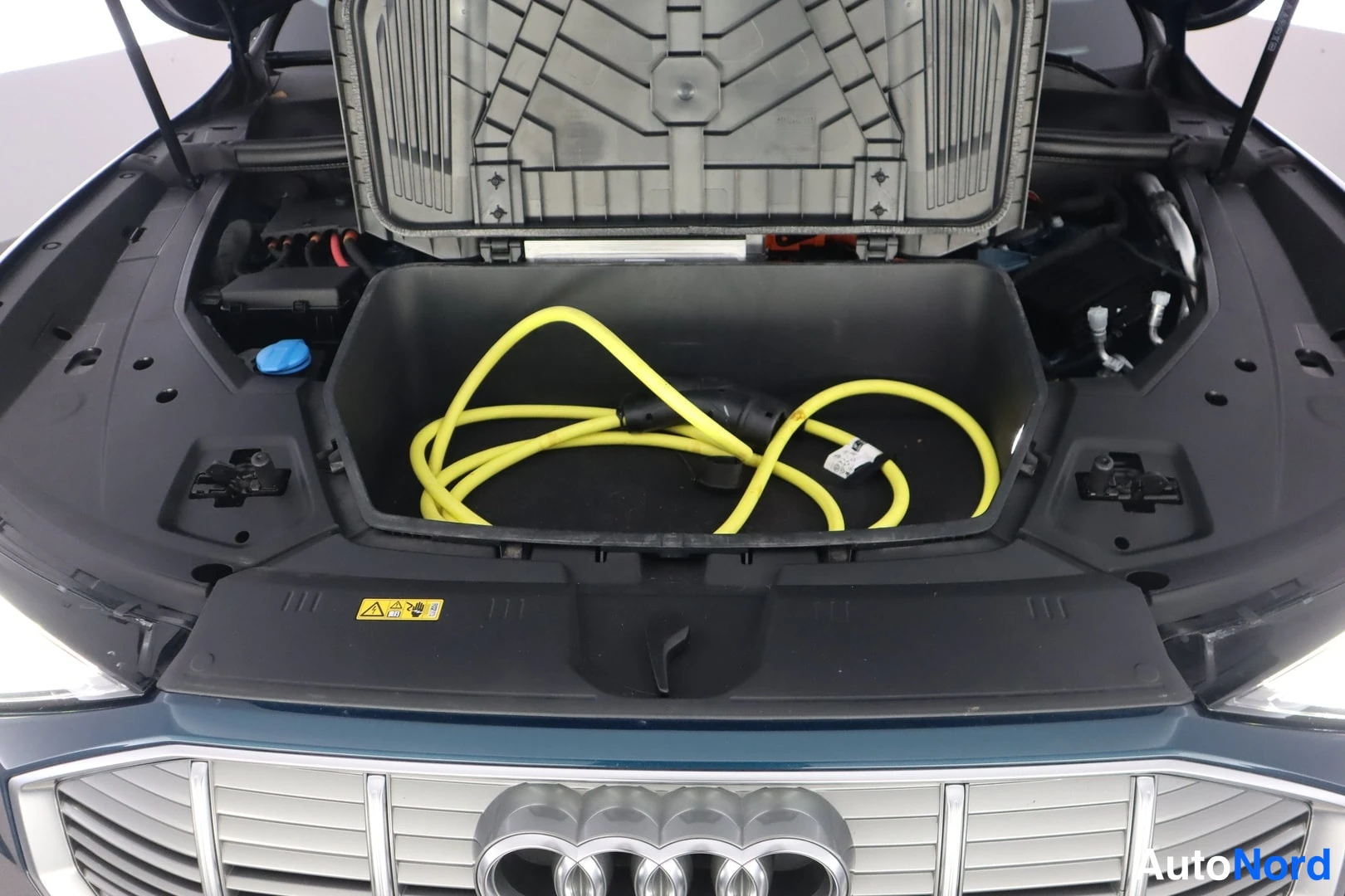 Edestä: Audi-merkillisen auton avattu etukonepellin alla oleva tavaratila (frunk), jossa säilytetään keltainen latauskaapeli; Audi-renkaat neljässa renkaassa konepellin