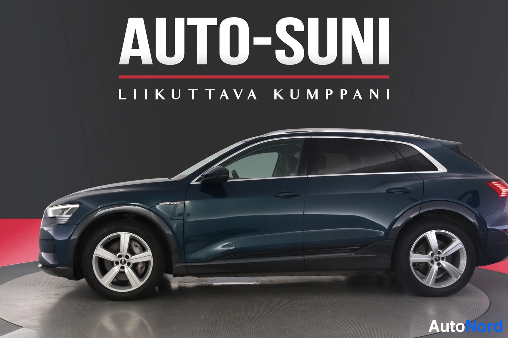 Sivulta: Tummansininen Audi e-tron -SUV kylkinäkymässä studiossa, taustalla tumma seinä ja puna-valkoinen Auto-Suni-logo.