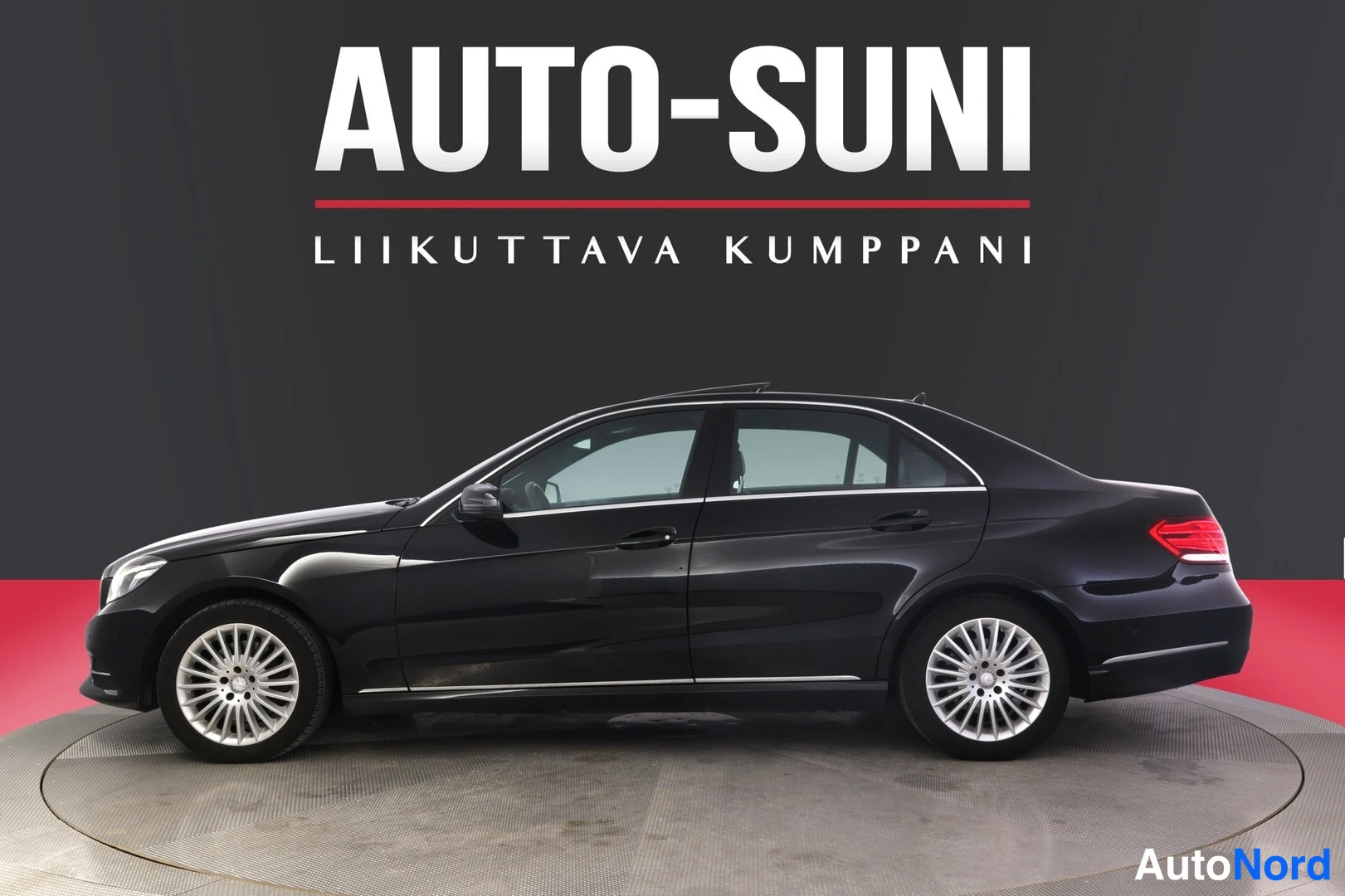 Sivulta: Mustan Mercedes-Benz E-luokan sedanin vasen kylki, jossa näkyvät monipuoliset kevytmetallivanteet, kattopaneelin lasiluukku ja takana punaiset takavalo