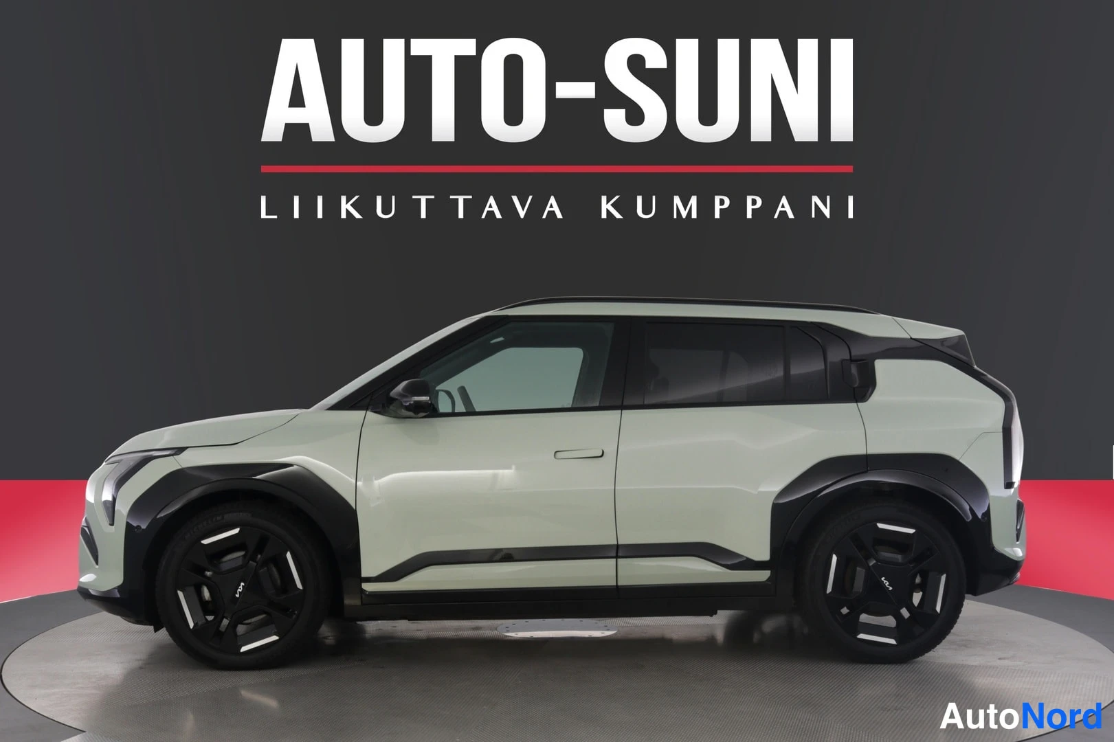 Sivulta: Vaaleanvihreä Kia EV3 crossoverelectricvehicle sivusta studiokuvassa, mustat kiiltävät kolot ja pyörät, tausta Auto-Suni-mainosgrafiikkaa