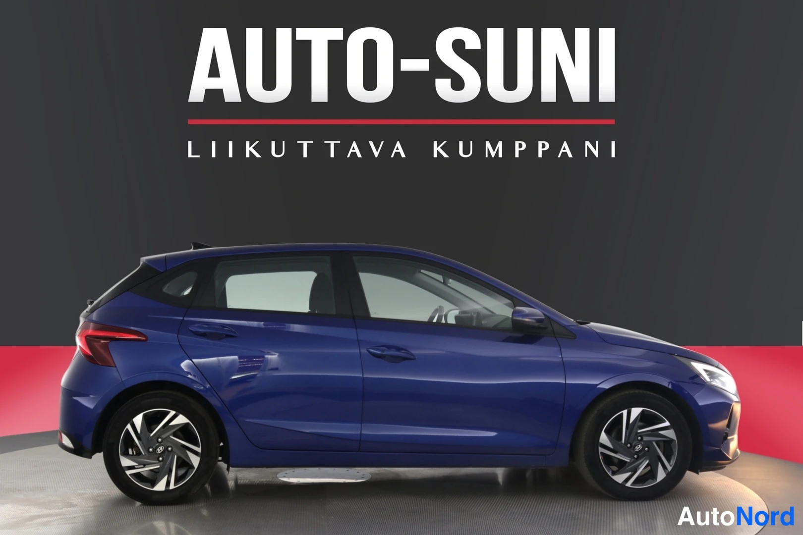 Etuviisto, nokka oikealle: Sininen Hatchback autotallissa, kokonainen auton vasen sivu ja etuosa näkyvät. Auto näyttää olevan hyvässä kunnossa. Taustalla on tumma seinä ja punainen