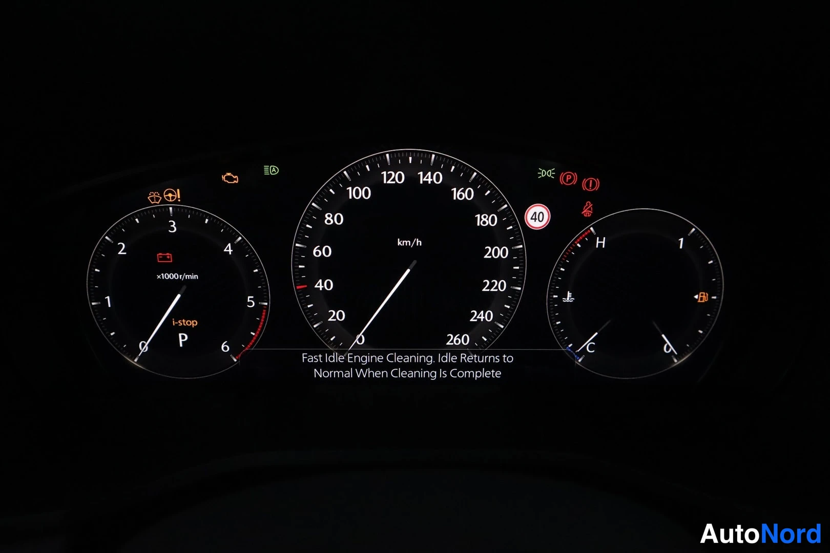 Sivulta: Mittaristo pimeässä: nopeusmittari asteikolla 0–260 km/h, kierrosmittari sekä polttoaine- ja lämpömittarit; näytöllä englanninkielinen teksti 'Fast Idle Engine