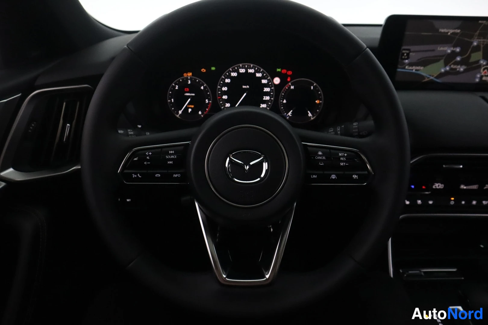 Sivulta: Ohjauspyörä Mazda-tunnuksella, sen takana analogiset mittarit ja oikealla infotainment-näyttö, joka näyttää navigointikarttaa.