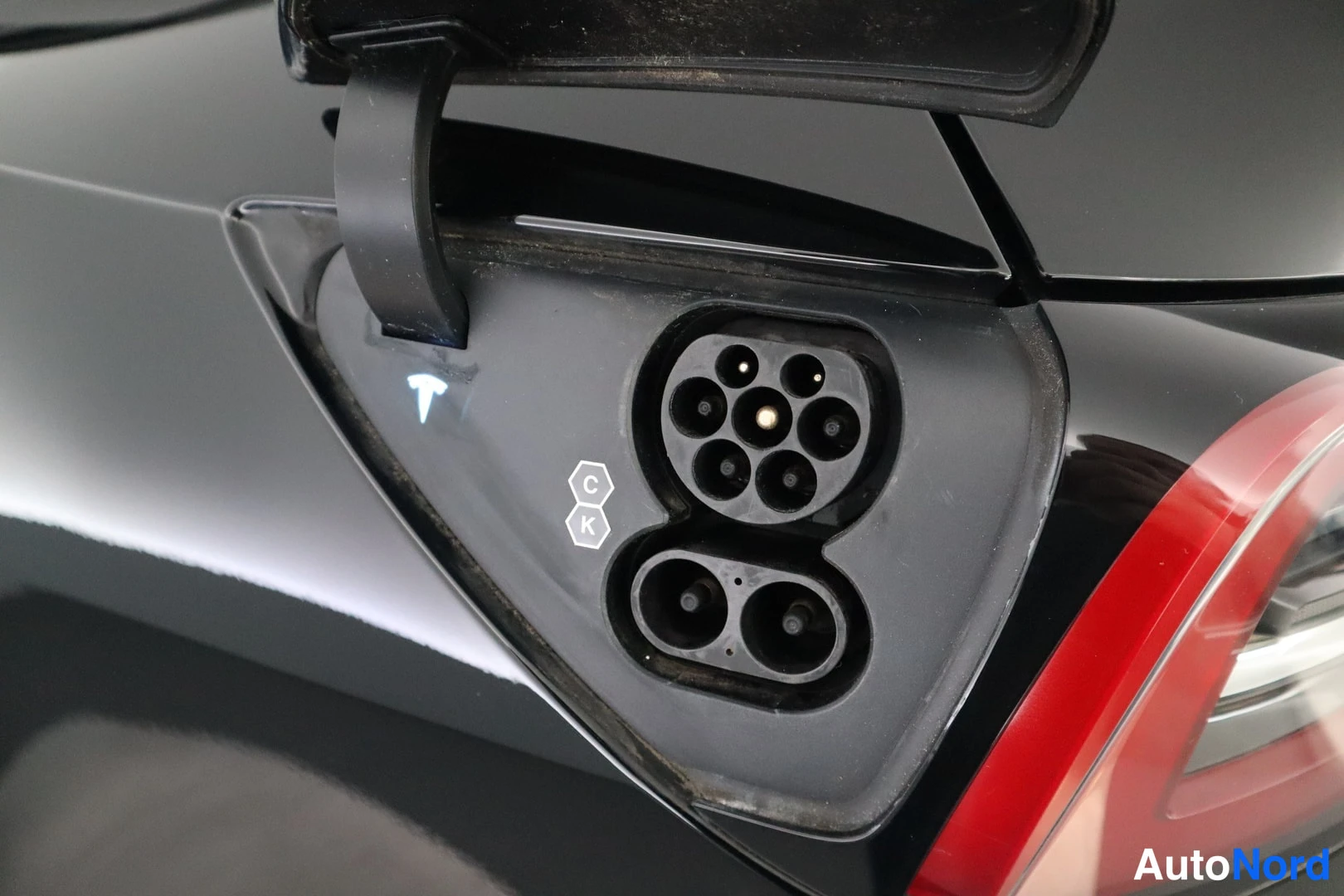 Yksityiskohta: Mustan auton latausportti auki: Type 2 -liitin, C/K-merkinnät ja Tesla-logo; vasemmalla punainen takavalo