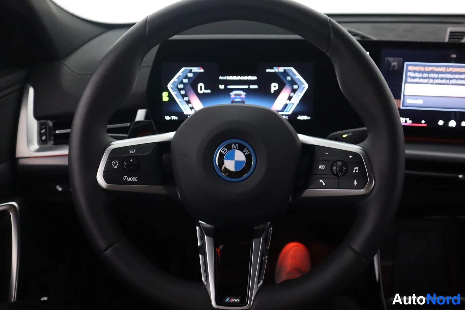 Muu: Ohjaamokuva: BMW-ohjauspyörä ja digitaalinen mittaristo; keskellä laaja näyttö, jonka teksti osittain epätarkka.