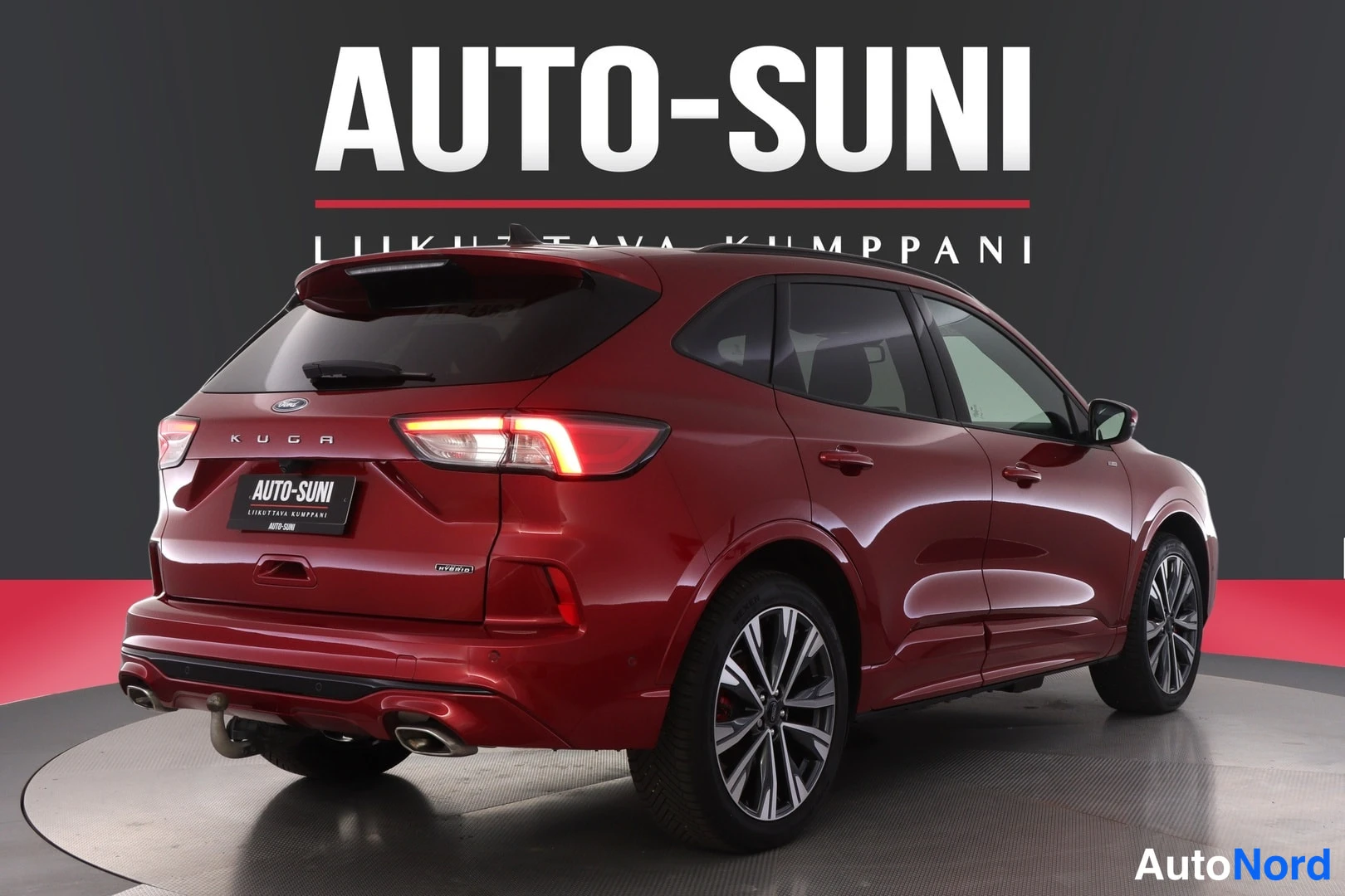 Takaviisto: Punainen Ford Kuga -henkilöauto takapäistä vasemmalta kuvattuna, takavalot päällä, AUTO-SUNI-mainosteksti taustalla.