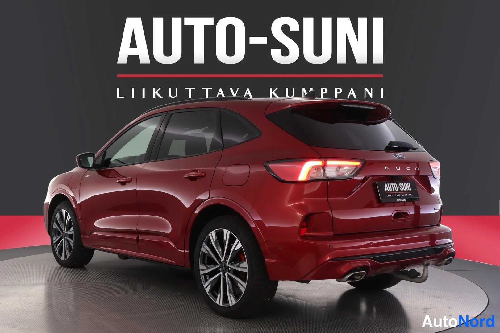 Takaviisto: Punainen Ford Kuga -henkilöauto takapäistä oikealta kuvattuna, takavalot päällä, AUTO-SUNI-mainosteksti taustalla.