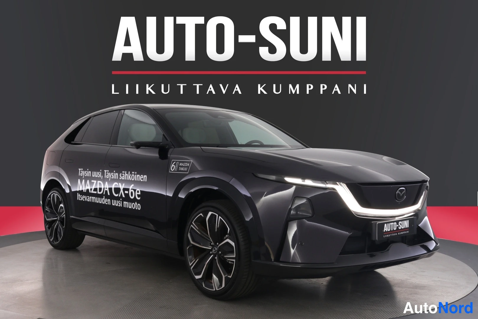 Etuviisto, nokka oikealle: Musta Mazda CX-6e kuvattu kolmasosa-etuviistosta oikealta nähtynä, Auto-Suni-kyltti keulassa ja mainosteksti kyljessä