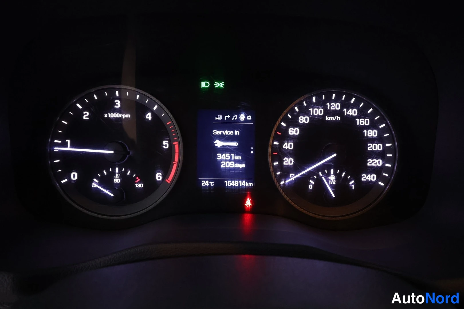 Yksityiskohta: Kojetaulu pimeässä: kierrosmittari ja nopeusmittari 0-240 km/h, keskellä näyttö Service In 3451 km 209 days ja kokonaismatka 164814 km.