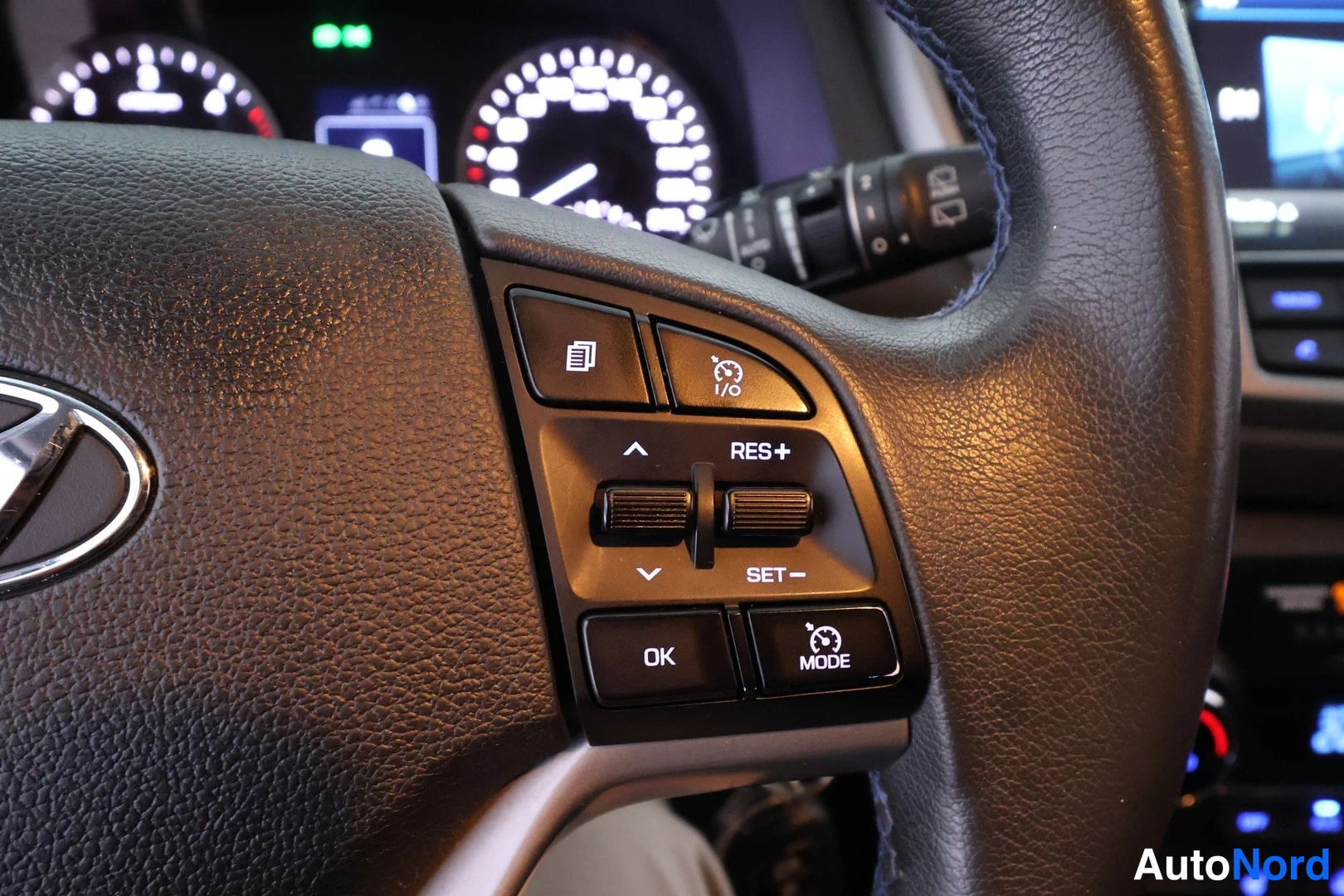 Yksityiskohta: Hyundai-ratin nappirivi lähikuvassa: RES+, SET-, OK, MODE ja I/O -napit, taustalla sumennettu kojetaulu.