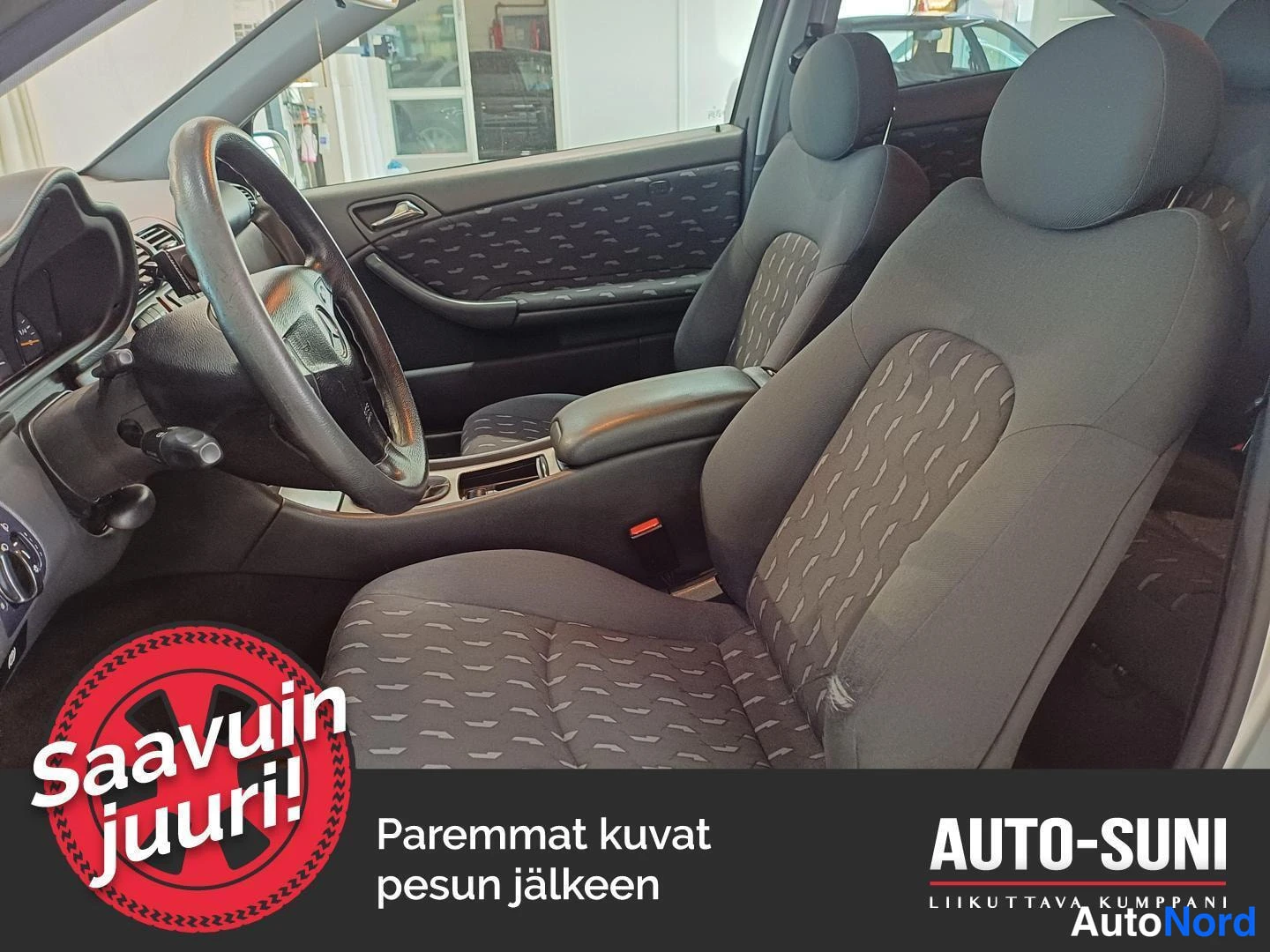 Etuistuimet: Interior view of the front seats and dashboard. Two front seats with grey fabric upholstery featuring patterned inserts are visible, along with the steering whe
