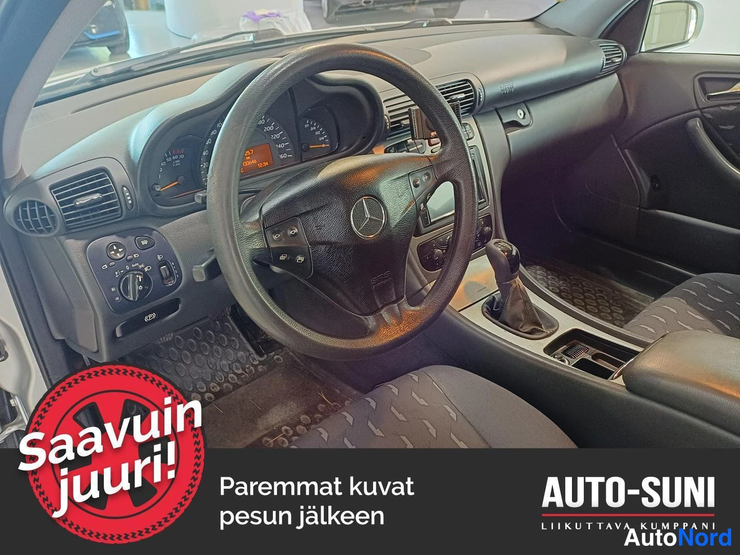 Etuistuimet: Dashboard, steering wheel and manual gear shift viewed from the driver's side. The instrument cluster is illuminated showing an orange display. To the left of t