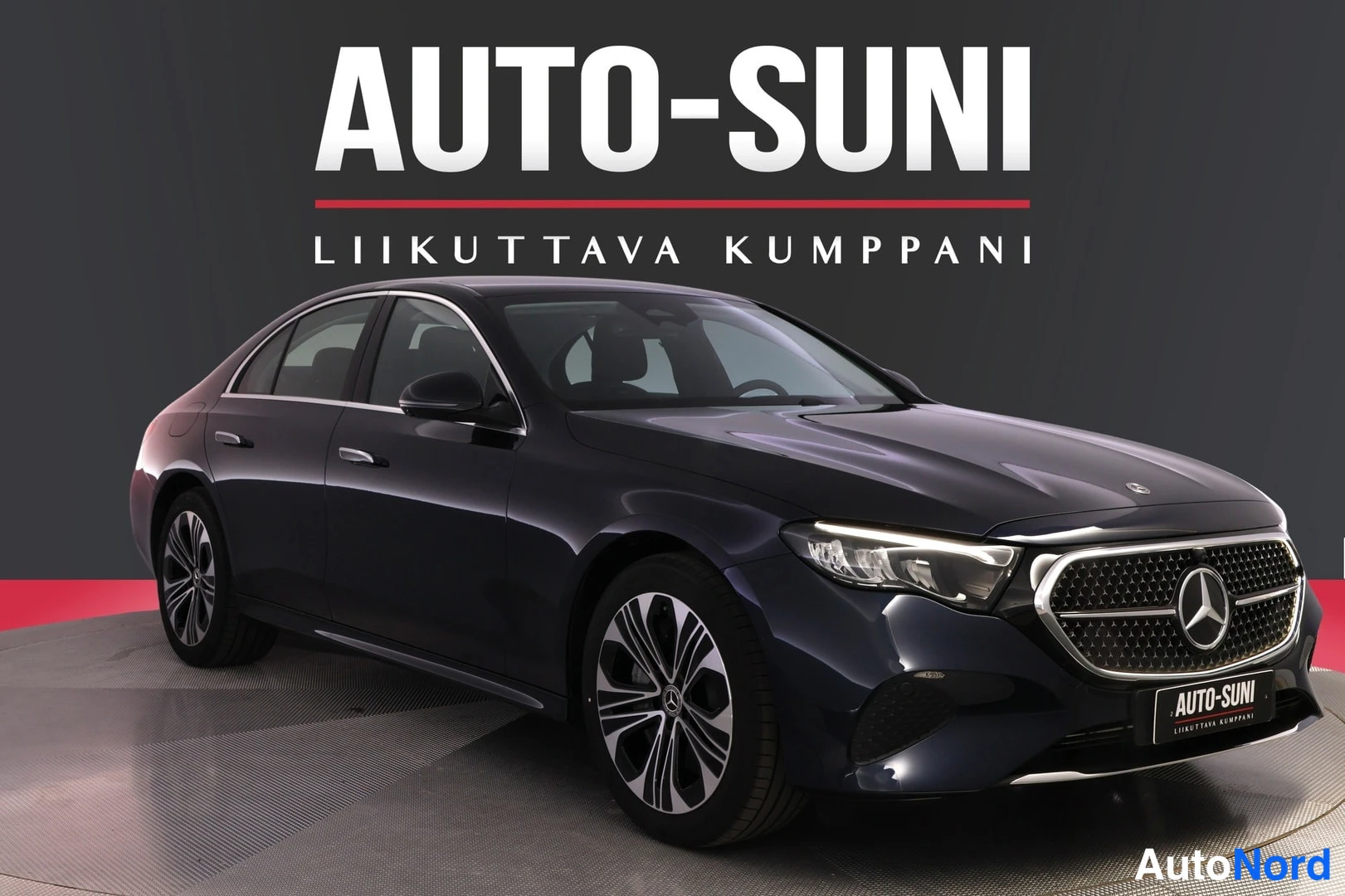 Etuviisto, nokka oikealle: Tummansininen Mercedes-Benz-sedan etuviistosta oikealta nähtynä valokuvastudiossa, AUTO-SUNI-logolla taustalla.