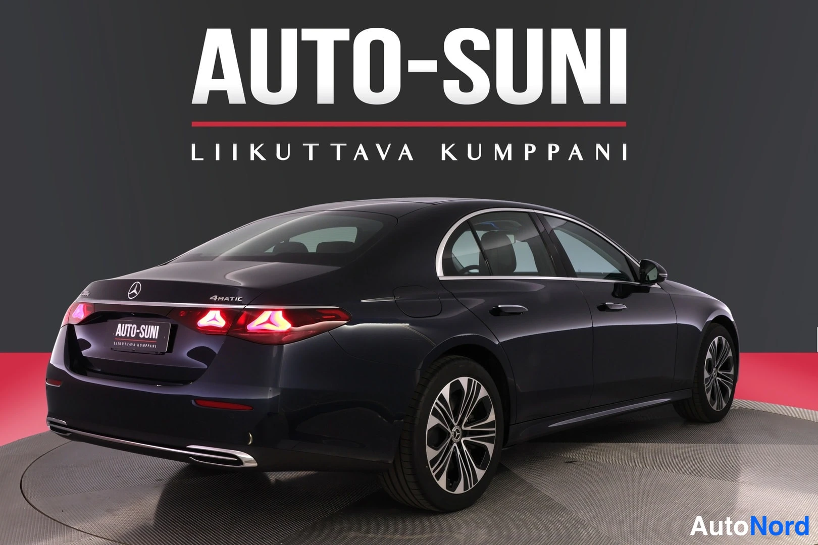 Takaviisto: Tummansininen sedan takaviistosta vasemmalta, sytytetyt takavalot, AUTO-SUNI-logo taustalla.