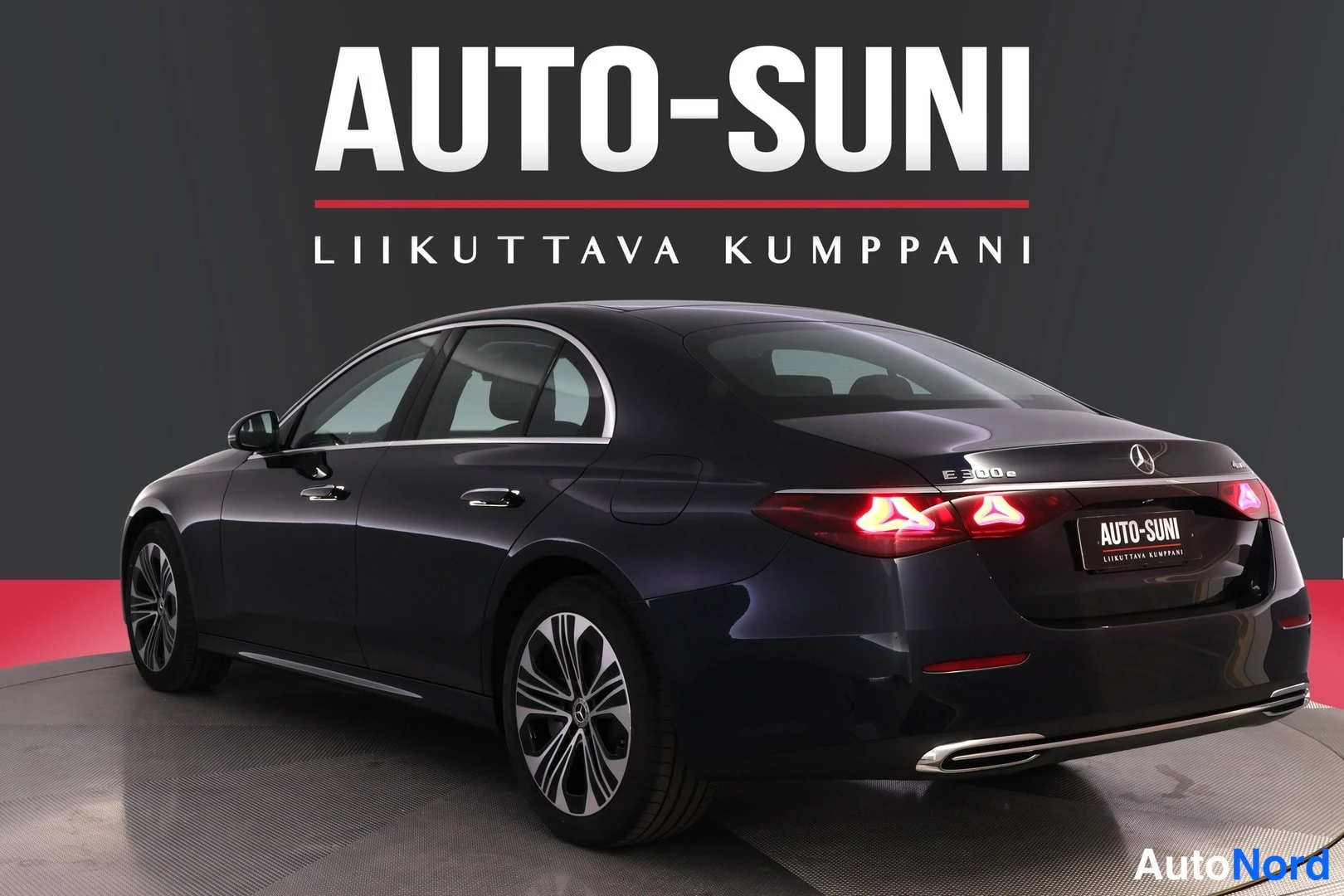 Takaviisto: Tummansininen sedan takaviistosta oikealta, sytytetyt takavalot ja pakoputket, AUTO-SUNI-logo taustalla.