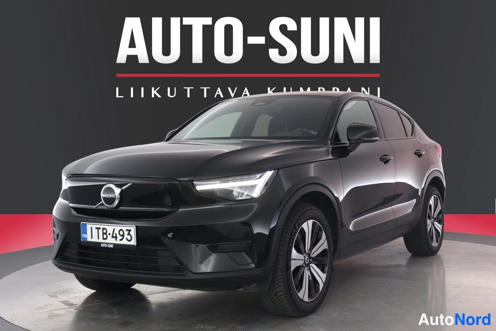 Etuviisto, nokka vasemmalle: Mustan auton etukulma lähes suoraan edestä kuvattuna, Volvo-merkki ja rekisterikilpi näkyvissä, Auto-Suni-logo taustalla.