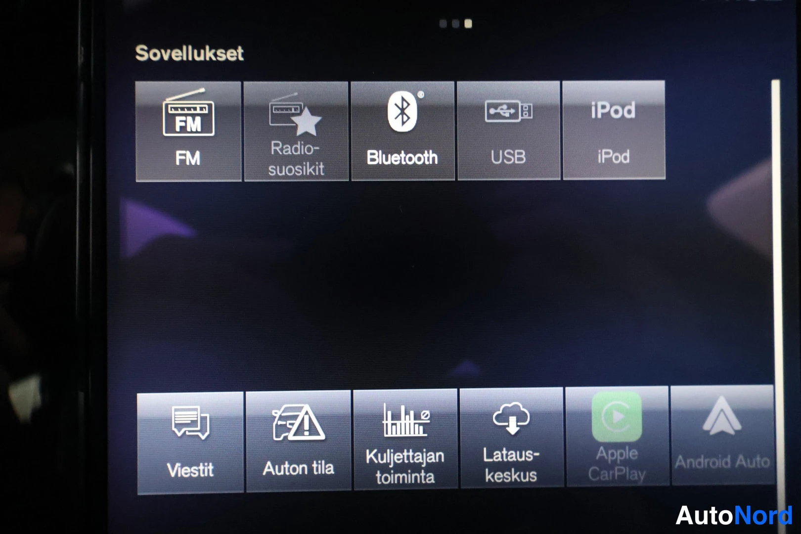 Muu: Auton näytön Sovellukset-valikko, jossa kuvakkeet FM, Radiosuosit, Bluetooth, USB, iPod, Viestit, Auton tila, Kuljettajan toiminta, Latauskeskus, Apple CarPlay