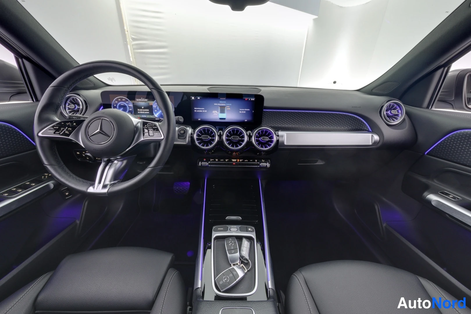 Ohjaamo: Interior cabin view showing multifunction steering wheel with two central screens, air vents and ambient lighting strips.
