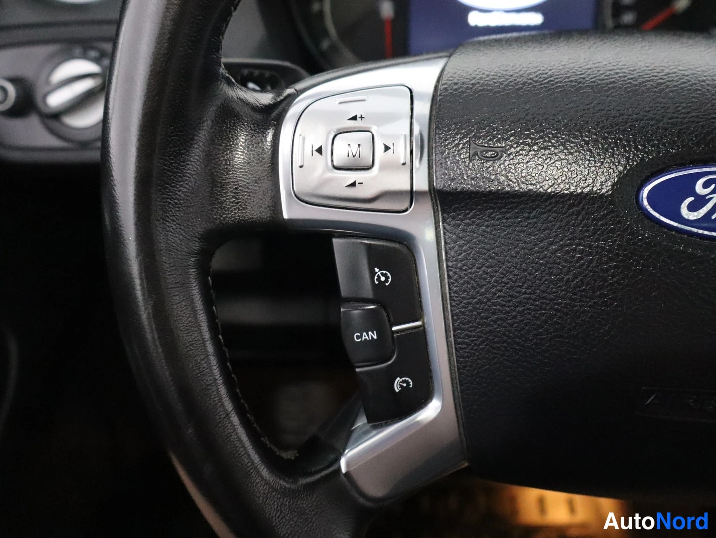 Edestä: Fordin ratin lähikuva näkyy vasemmalta. Ratissa on ääniohjaimet (M, +/-, CAN) ja sininen Ford-tunnus. Ratin kulmassa näkyy korni ja ilmastointipaneelin kääntöna