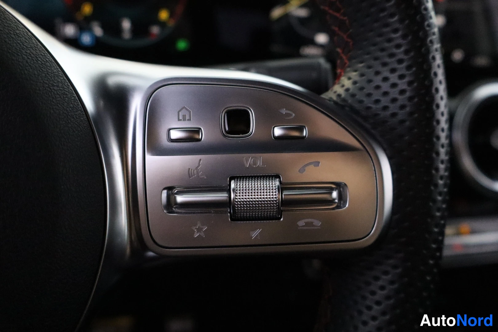 Yksityiskohta: Close-up of the left side of a perforated leather-wrapped multifunction steering wheel, showing control buttons for media (VOL), navigation, phone, and voice co