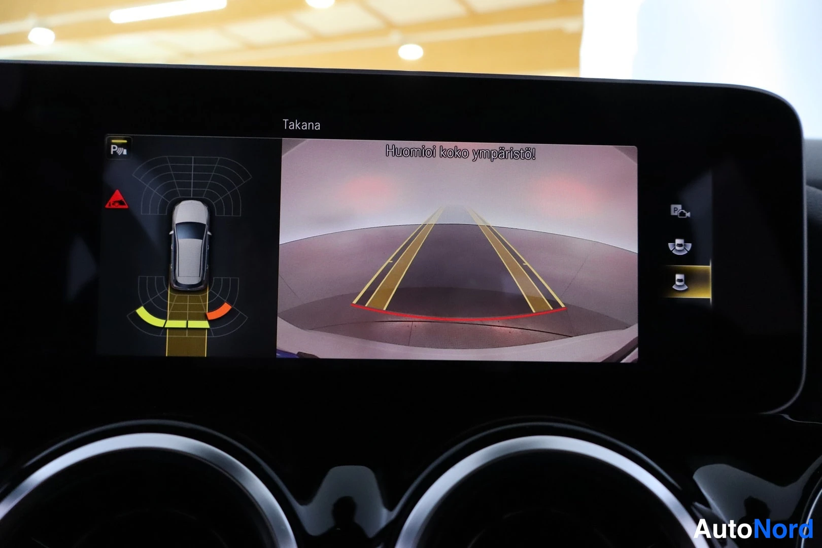 Ohjaamo: Central infotainment display showing the reverse camera view with guidance lines and a top-down graphical representation of the vehicle alongside parking distan