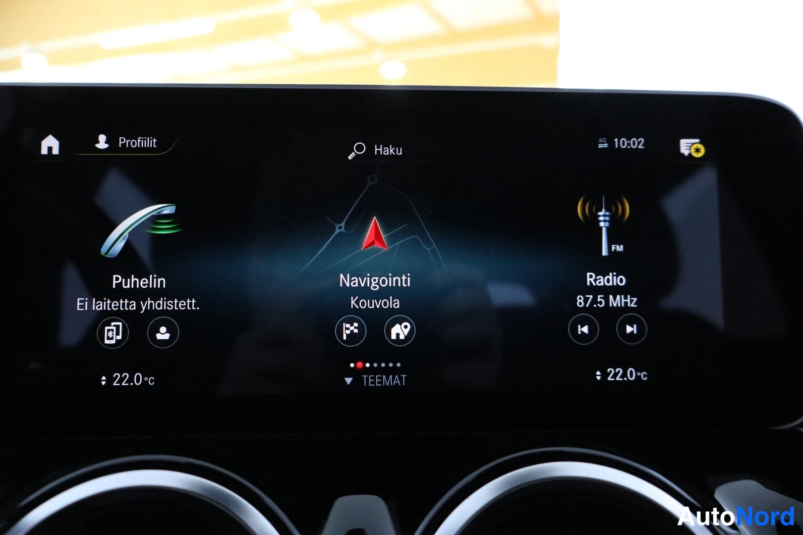Ohjaamo: Infotainment screen showing main menu options: 'Puhelin' (Phone), 'Navigointi' (Navigation) to Kouvolia, and 'Radio' set to 87.5 MHz. Time is 10:02.