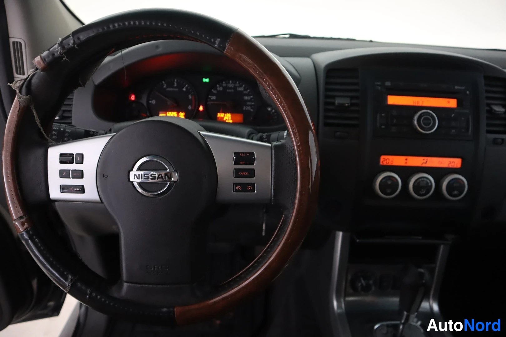 Muu: Ohjauspyörä Nissan-logolla, ruskeat puuvanteet, kojelaudan mittarit ja keskikonsolin radio sekä ilmastointipaneeli näkyvät sisätiloista otetussa kuvassa.