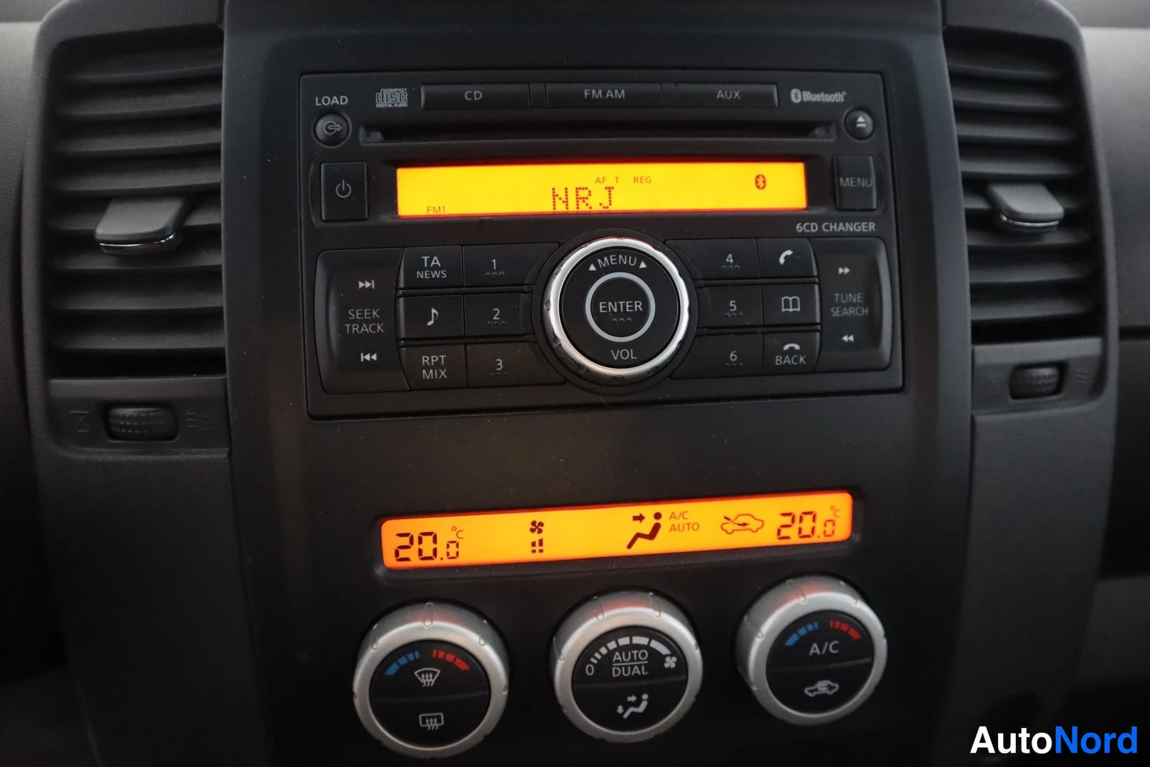 Muu: Keskikonsolin radio ja ilmastointipaneeli lähikuvassa: Bluetooth-merkintä, näytössä NRJ ja molempien puolen lämpötila 20°C, AUTO DUAL -painikkeet.
