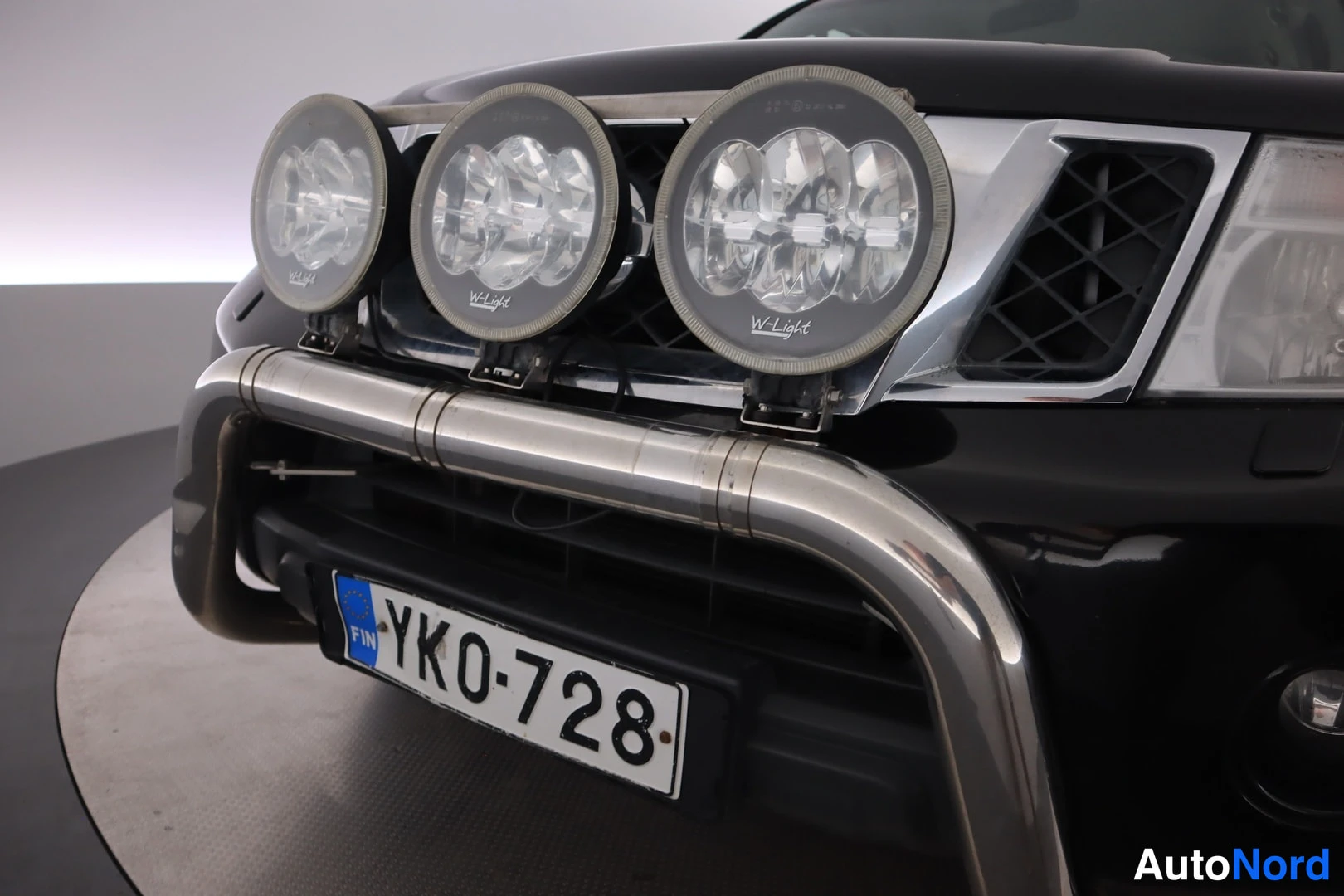 Edestä: Mustan auton etuosan lähikuva: etupuskuriin kiinnitetty teräskbögelissä kolme W-Light-ajovaloa, rekisterikilpi YK0-728.