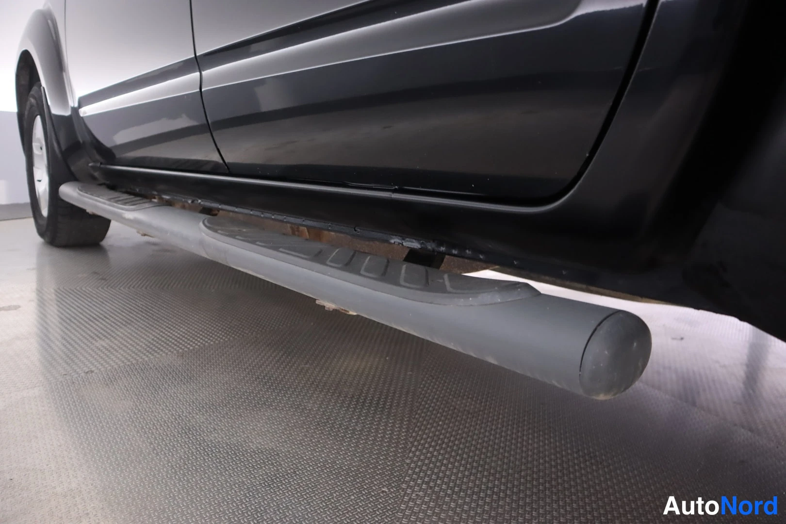 Sivulta: Lower side view of black car showing grey tubular side step with rubber tread pads along the sill.