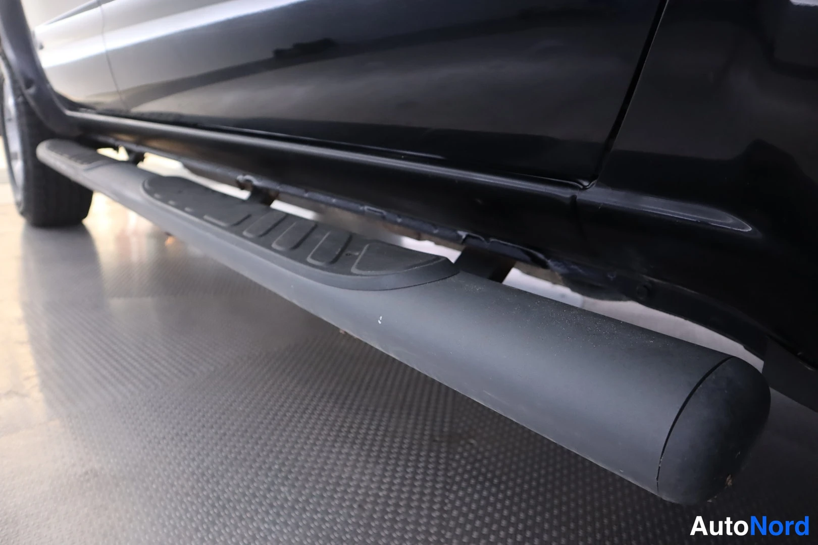 Sivulta: Close-up of grey tubular side step with black tread pad mounted on the sill of a black car.