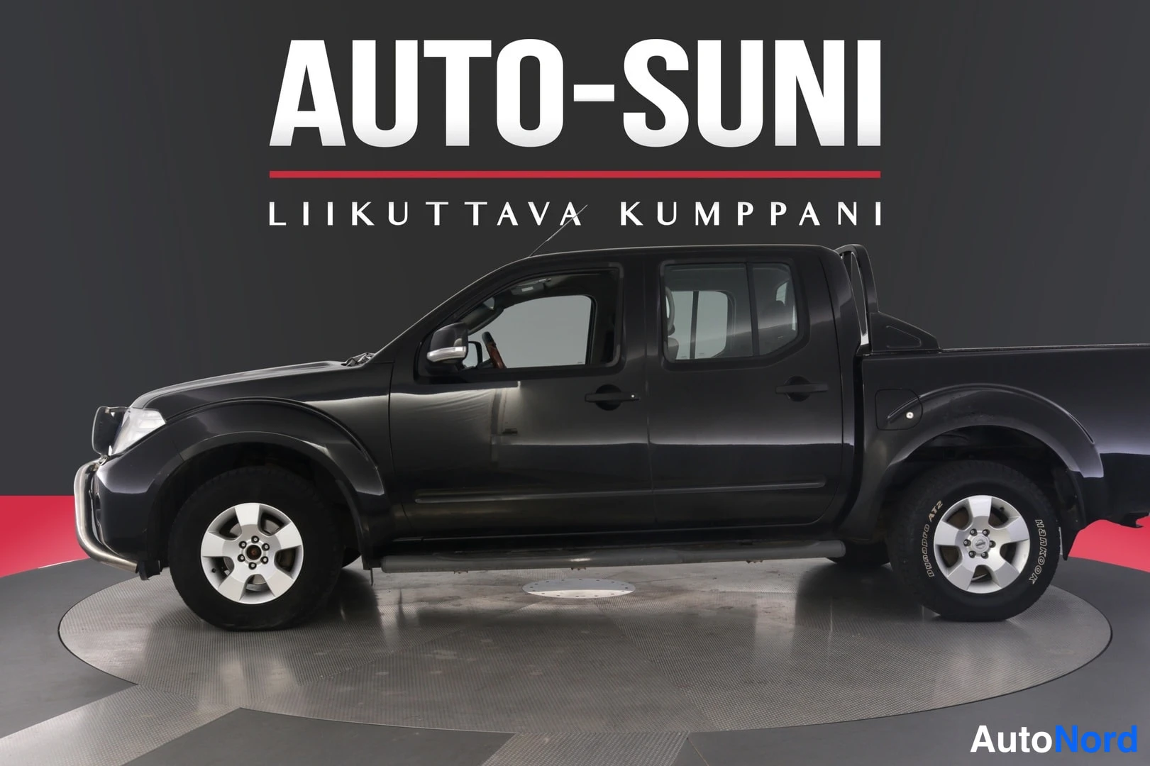 Sivulta: Musta Nissan Navara -mallinen avolava-auto sivulta etukolmannesta, nokka vasemmalla, studio-taustalla AUTO-SUNI-logolla.