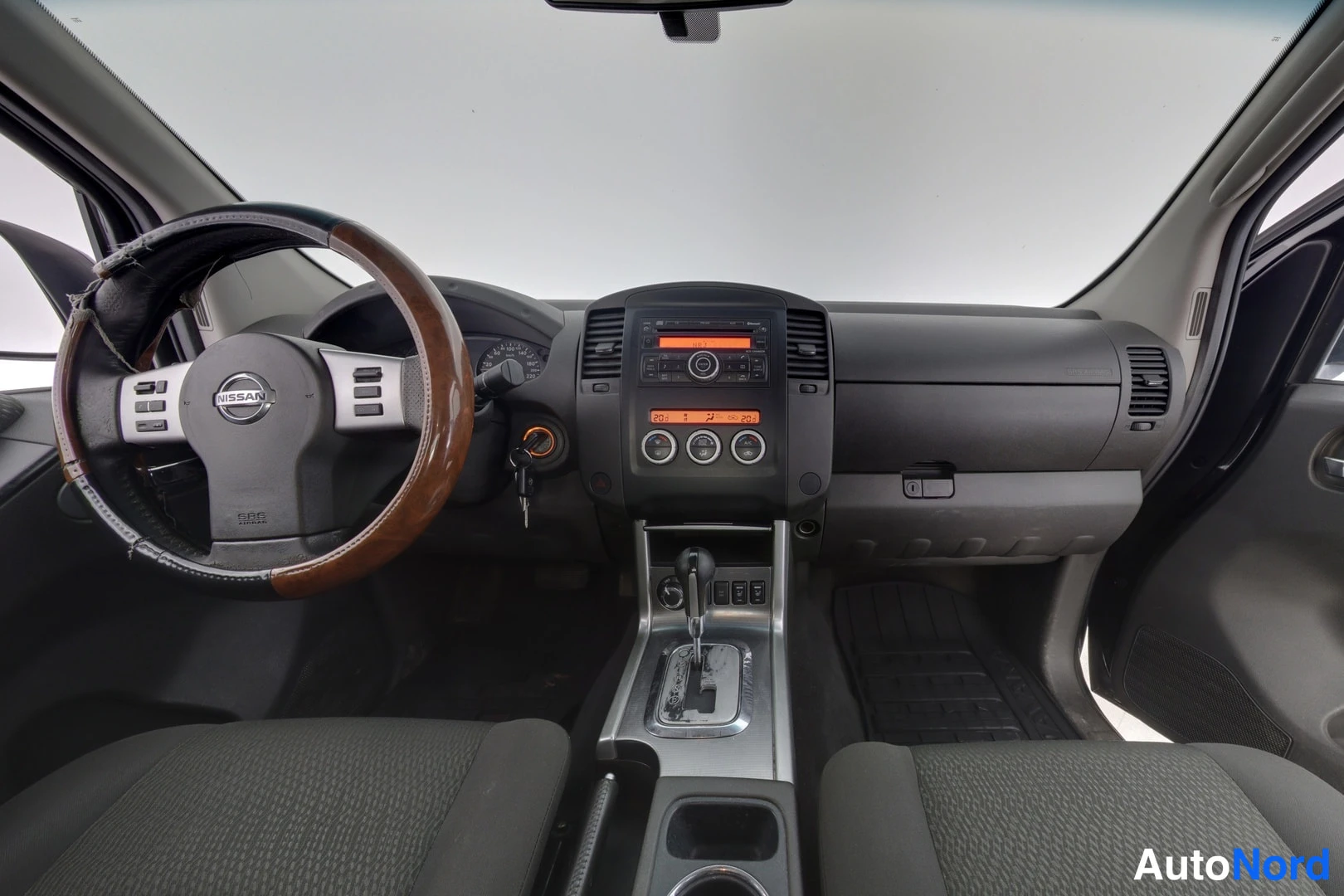 Yksityiskohta: Nissan Navaran ohjaamo: Nissan-merkitty ohjauspyörä puukuvioisella pinnalla, automaattivaihteiston valitsin ja keskusprojan radio.