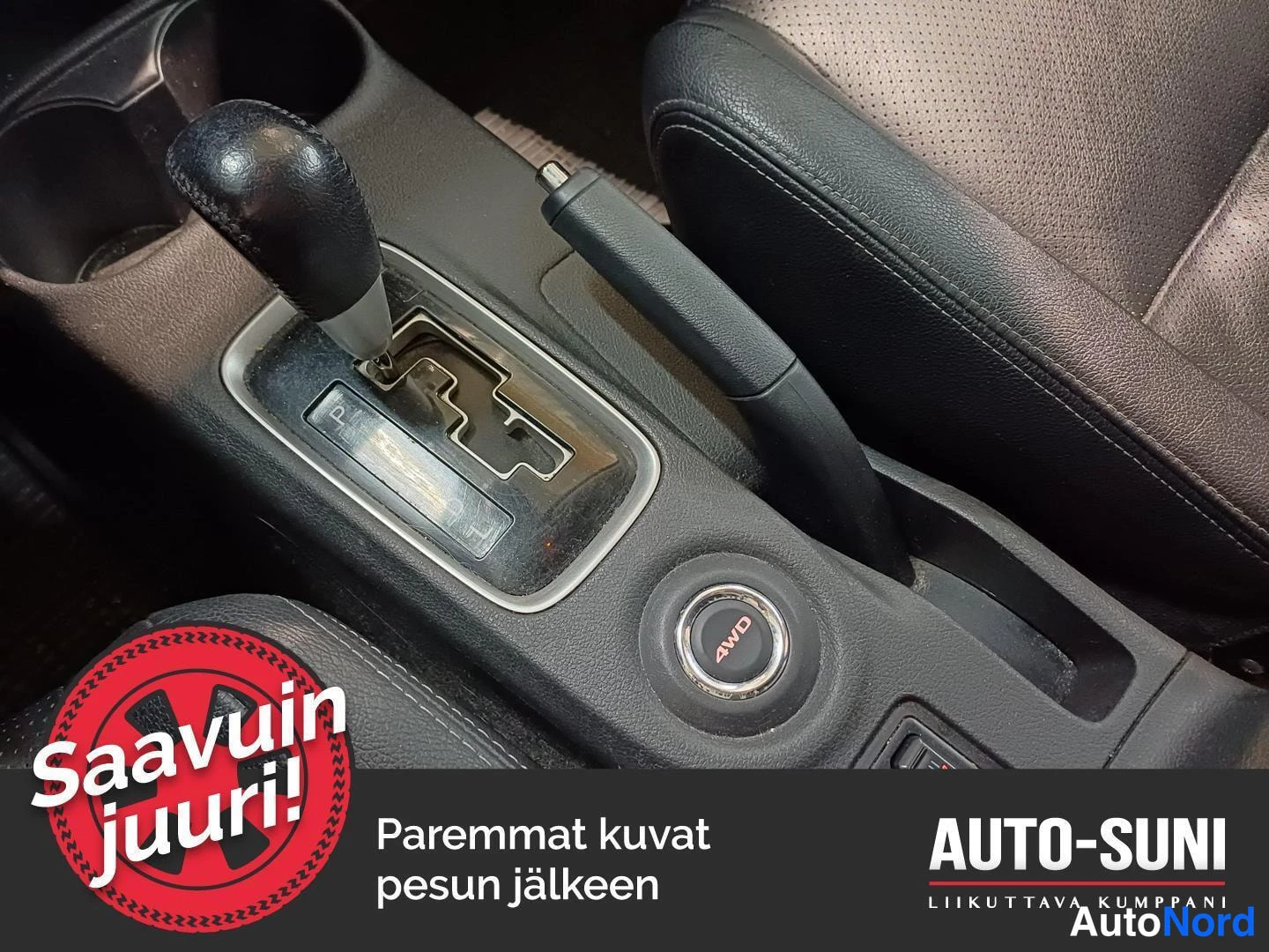 Yksityiskohta: Mitsubishi Outlanderin keskikonsolin vaihteenvalitsin, käsijarruvipu ja pyöreä AWD-nappi, jonka ympärillä harmaa kierto. Ohjaamon pinnat ovat tummia. Kuvassa on