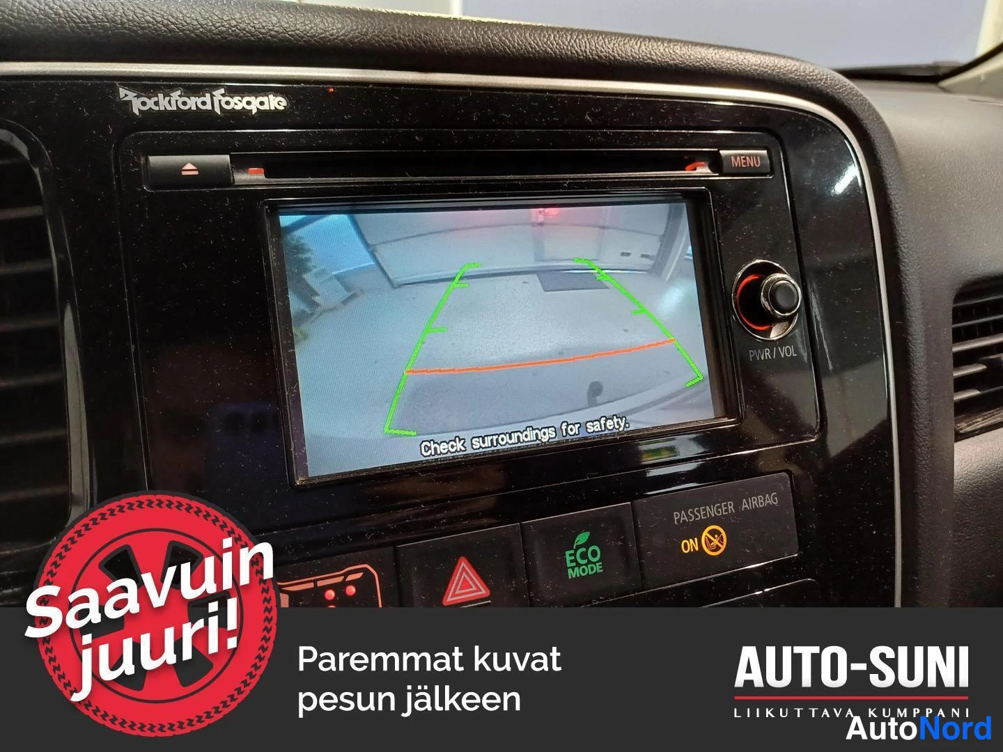Yksityiskohta: Mitsubishi Outlanderin näyttö, joka näyttää takakameran kuvan vihreällä ja oranssilla kaistaohjauksella sekä tekstin 'Check surroundings for safety.' Paneelissa